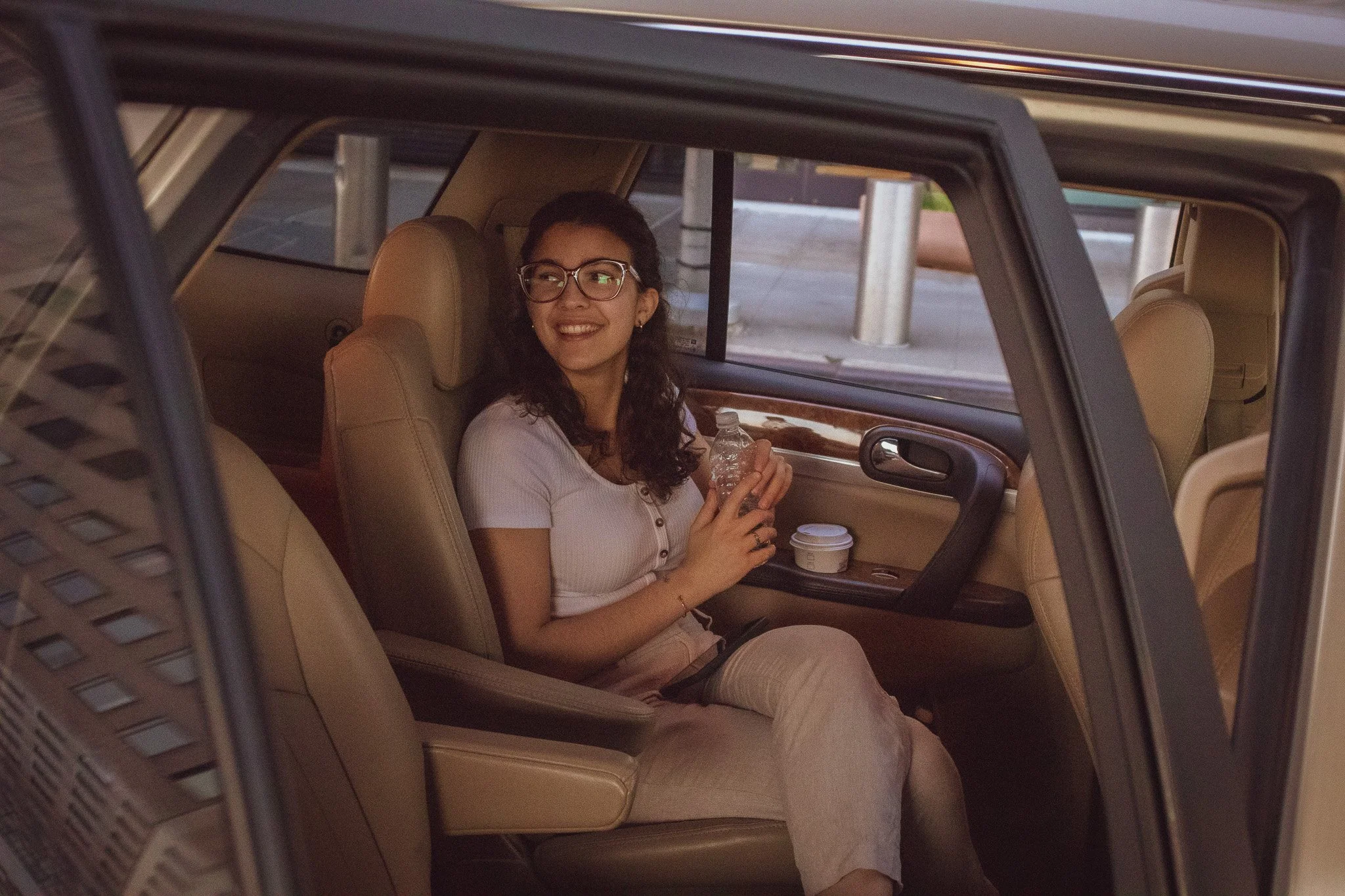A woman with glasses and dark curly hair sitting inside a car, smiling while holding a water bottle, with coffee cups on her lap.