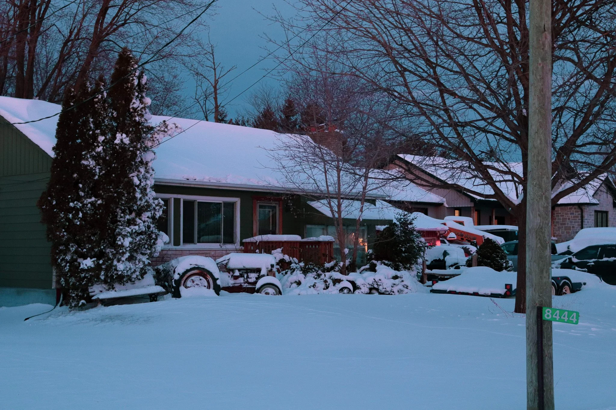 A snowy residential backyard scene with a house, leafless trees, snow-covered yard, and vehicles.