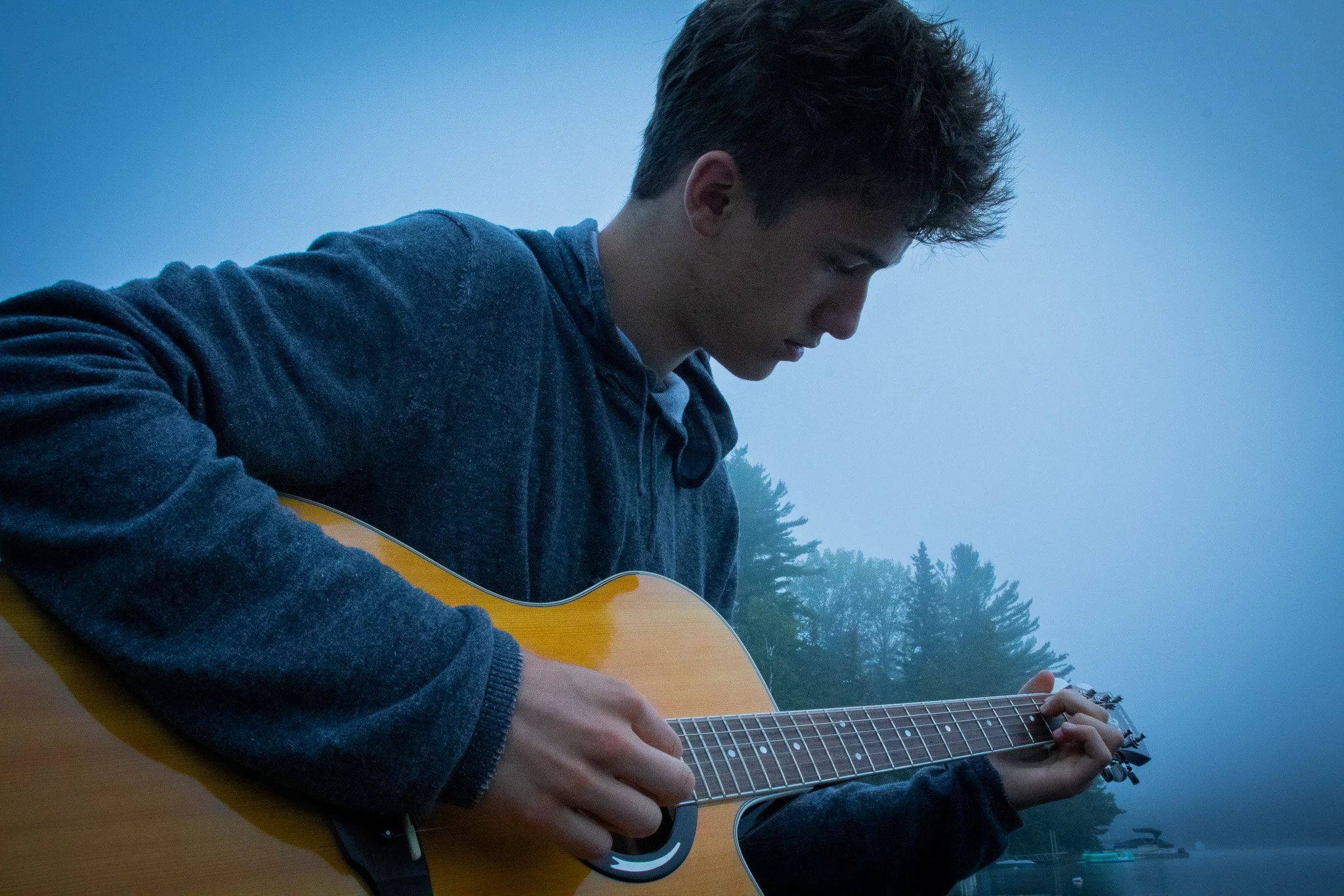 A young man with dark hair playing an acoustic guitar outdoors during foggy weather, with trees in the background.