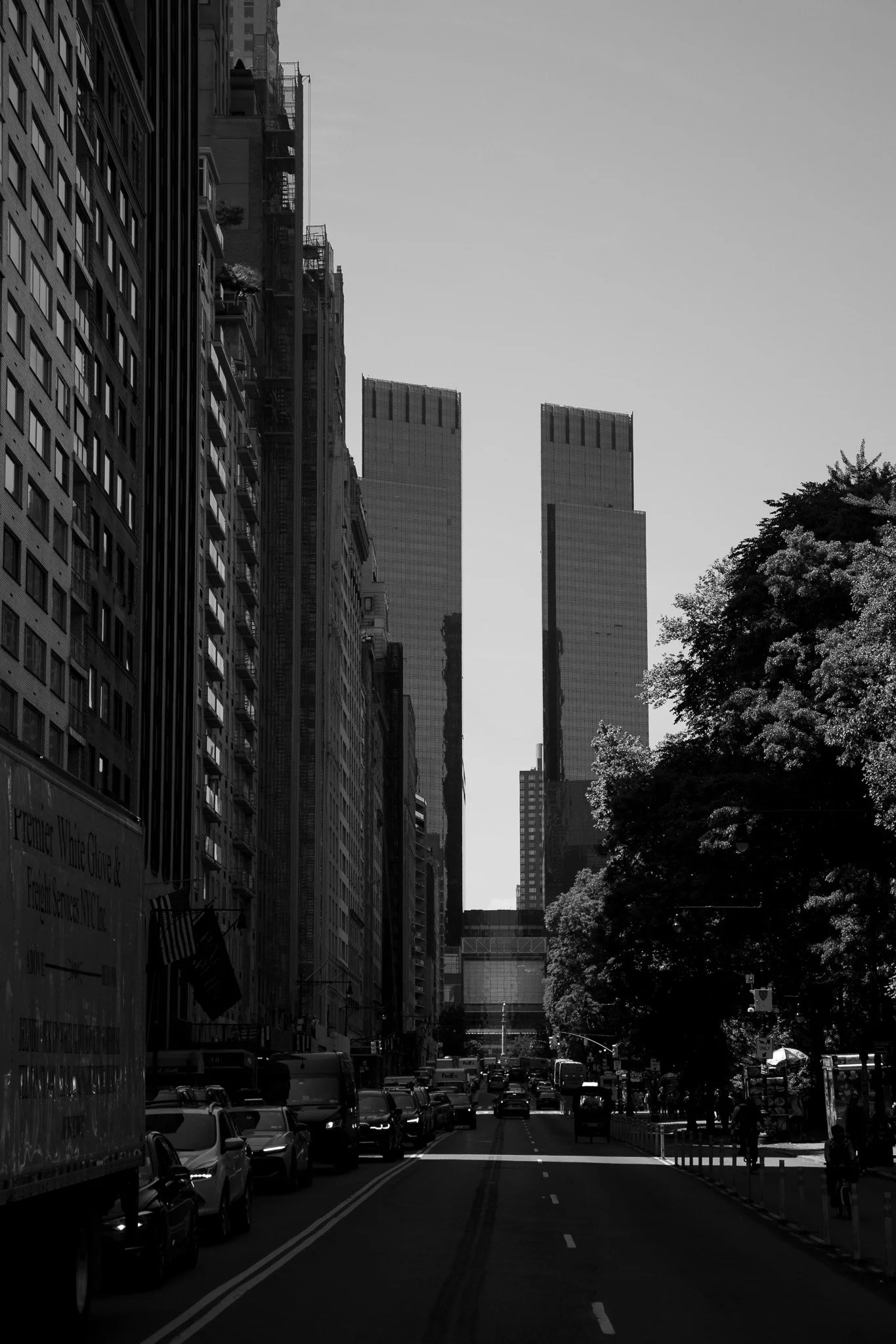 Black and white street scene in a city with tall buildings, cars, and trees lining the street.