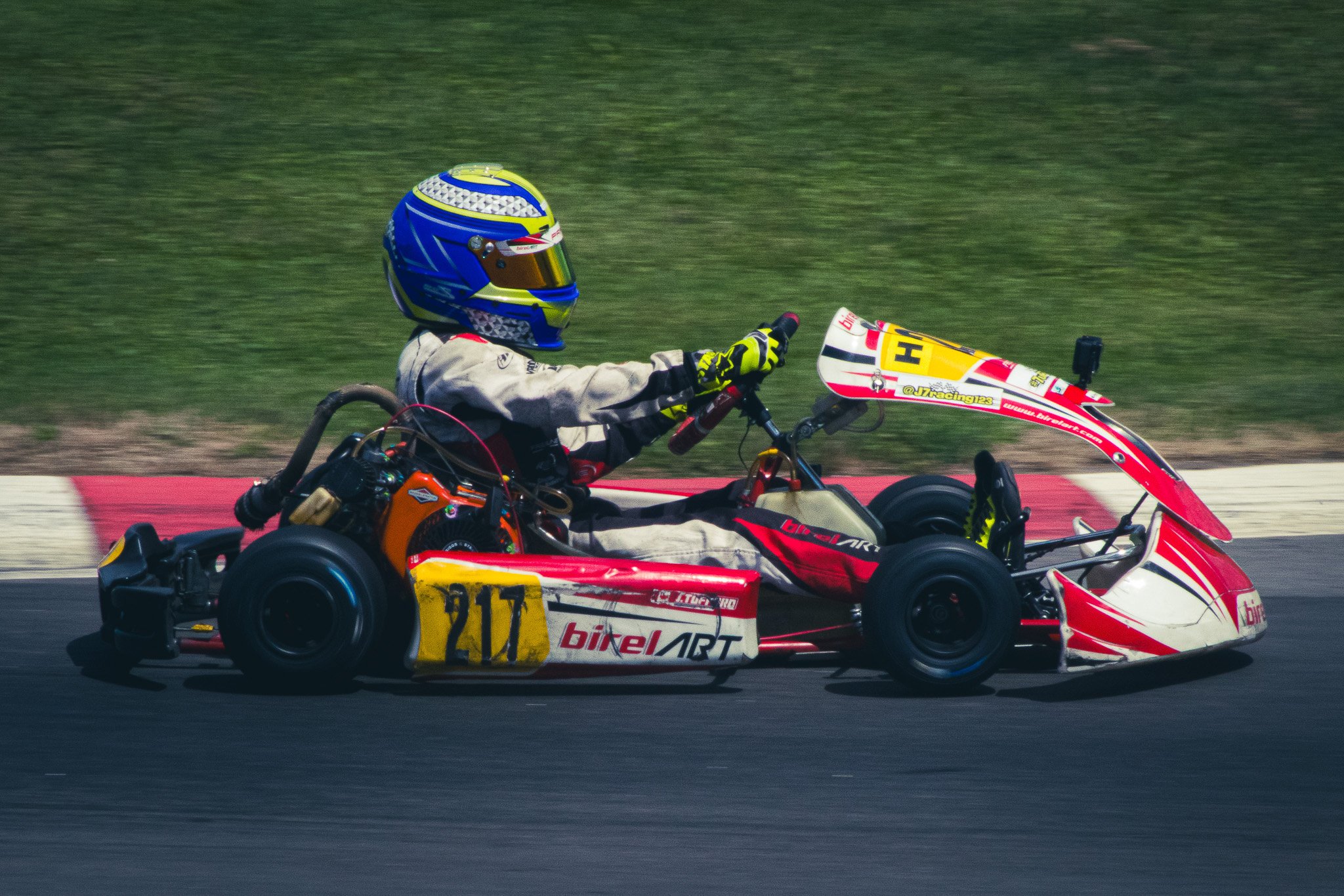 A go-kart with the number 211 racing on a track during daytime, driven by a person wearing a helmet and racing suit.
