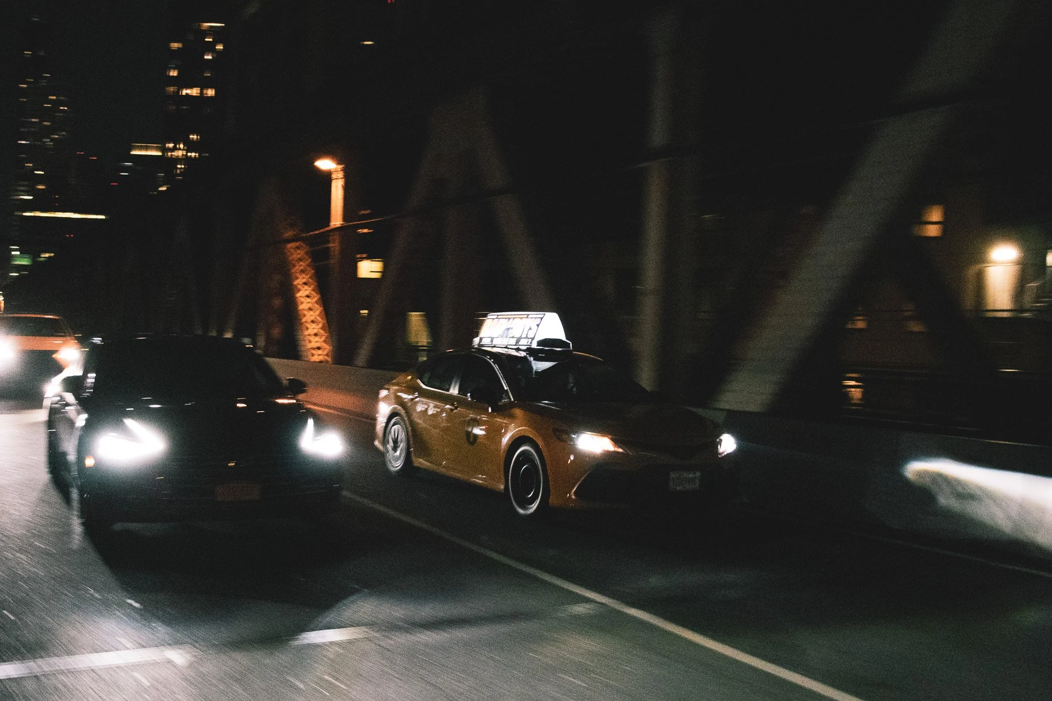 Nighttime scene of traffic on a city bridge with multiple cars, some headlights on, a lit streetlamp, and a building with illuminated windows in the background.