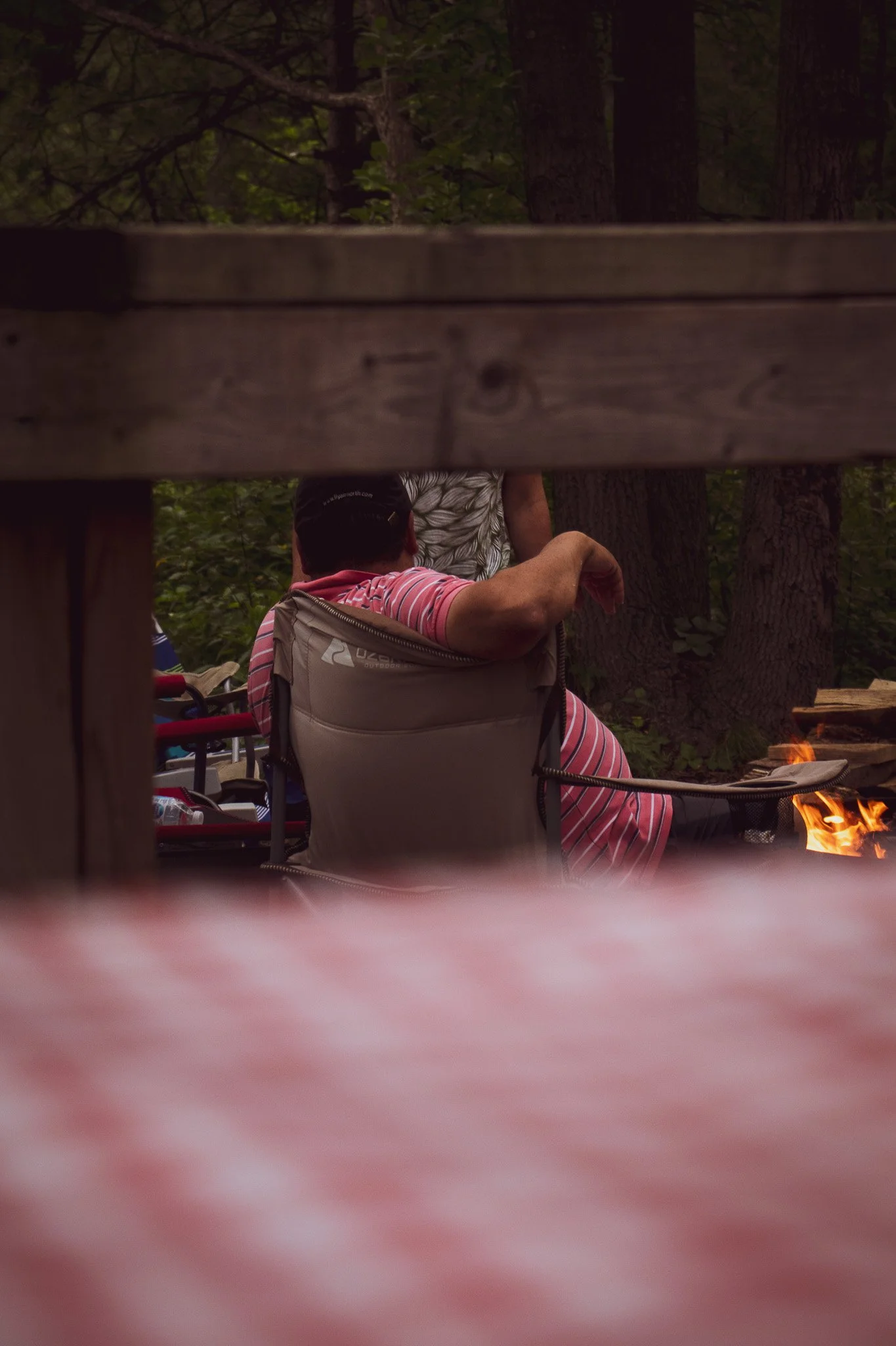 View from a wooden structure looking through the gap at a person seated outdoors, surrounded by trees, with a small fire nearby.