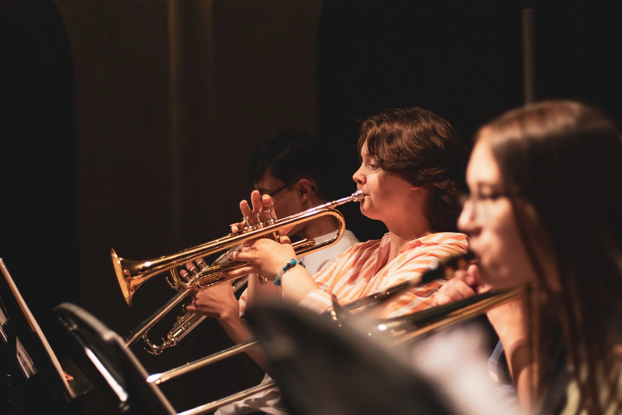 A group of people playing the trumpet in an orchestra, focusing on a woman in the center with a striped top.