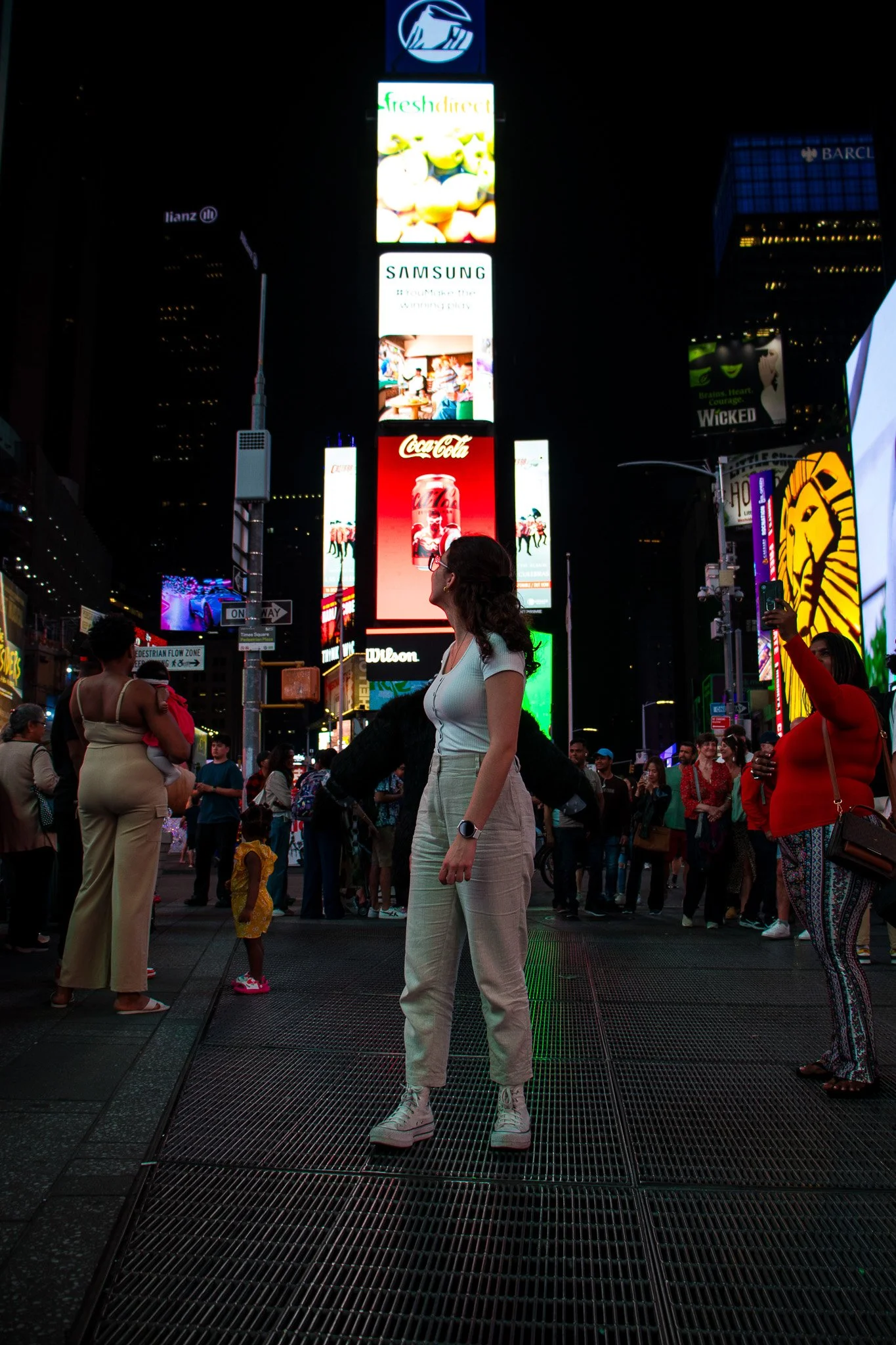 City crowd at night in Times Square with bright billboards and advertisements, including Coca-Cola, Samsung, and others, with people standing and walking around.