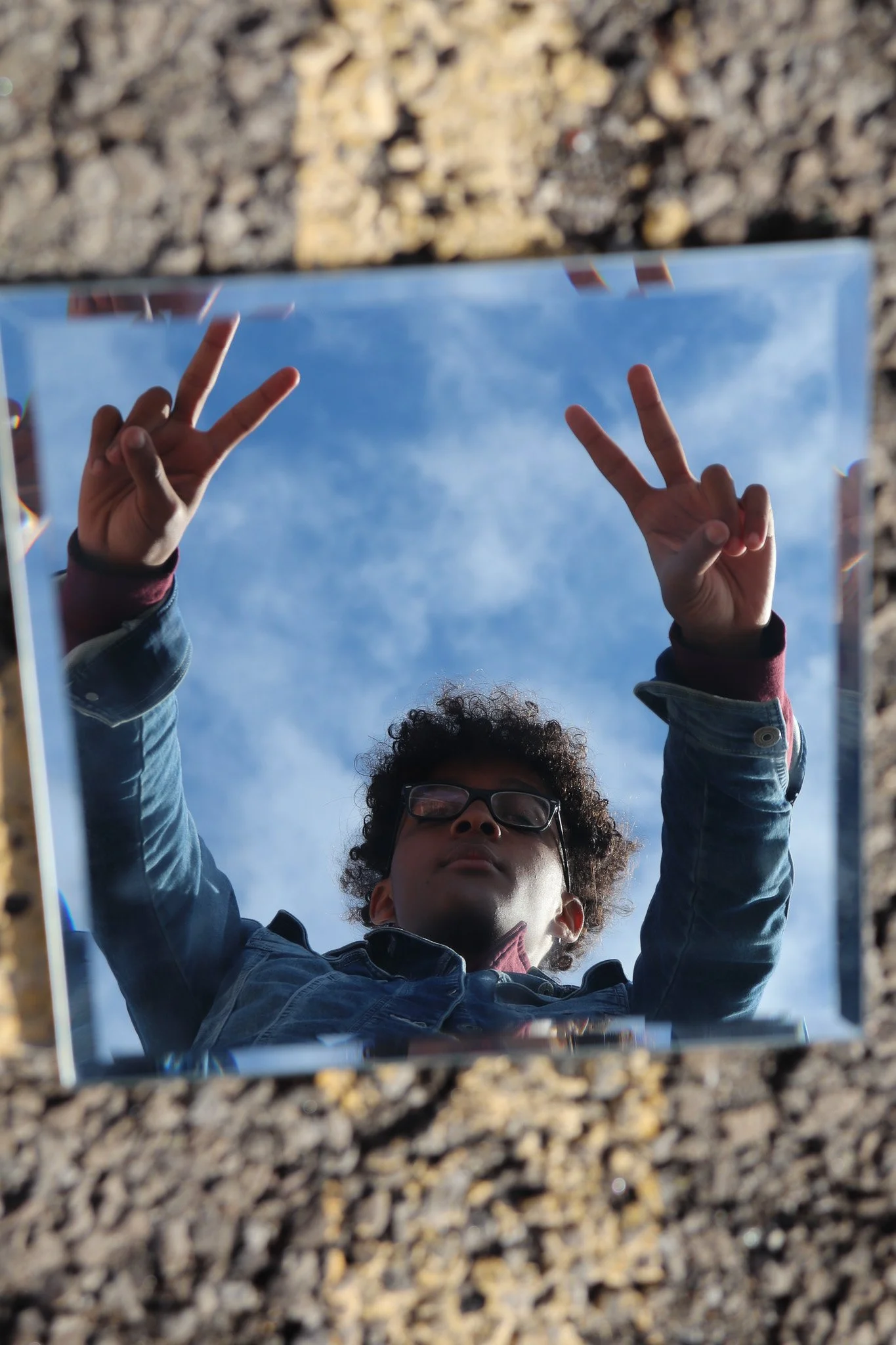 A person with curly hair and glasses taking a selfie with a mirror, making peace signs with both hands, against a blue sky background.