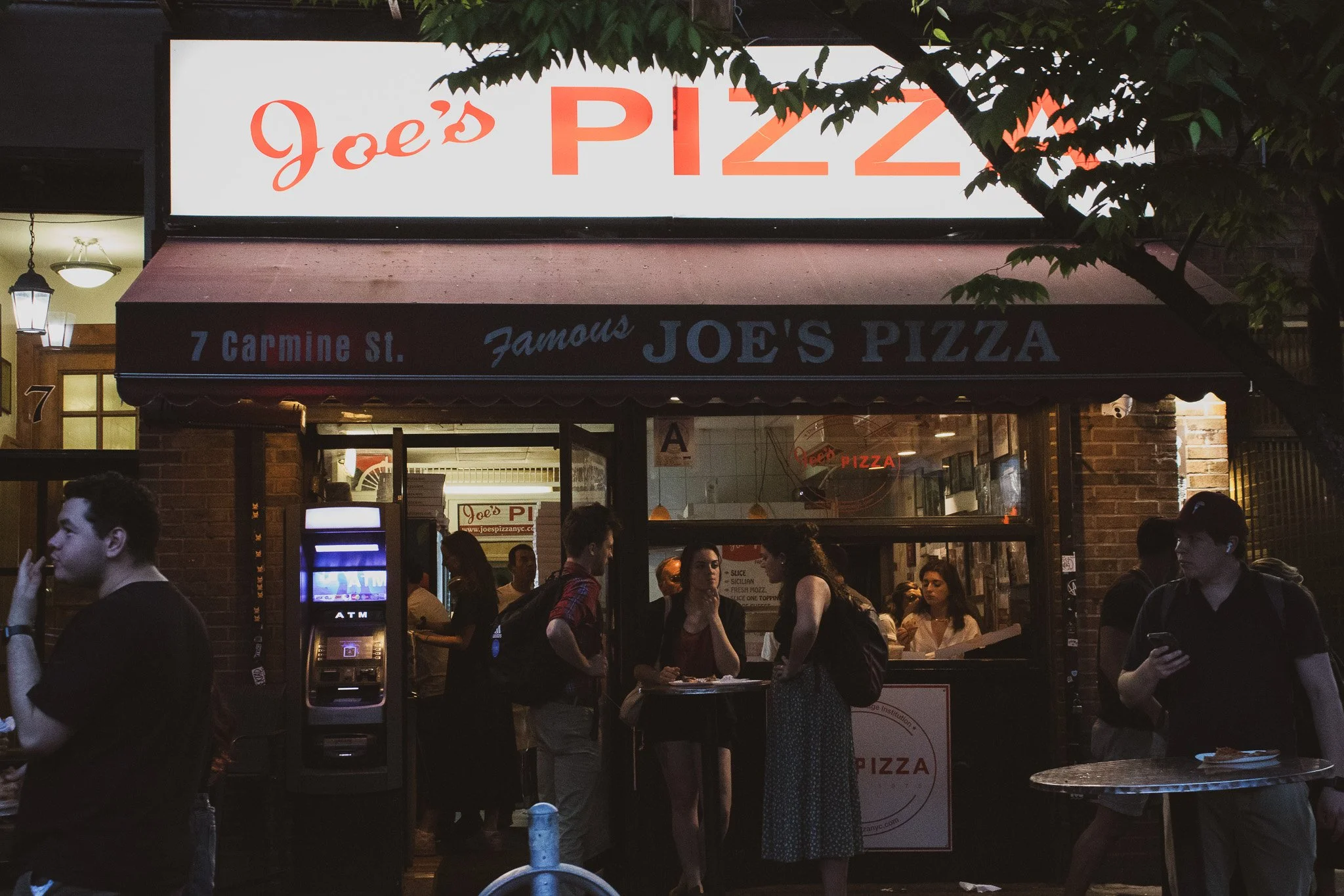Night view of Joe's Pizza restaurant with a bright sign, people gathered outside, and an ATM machine in front.