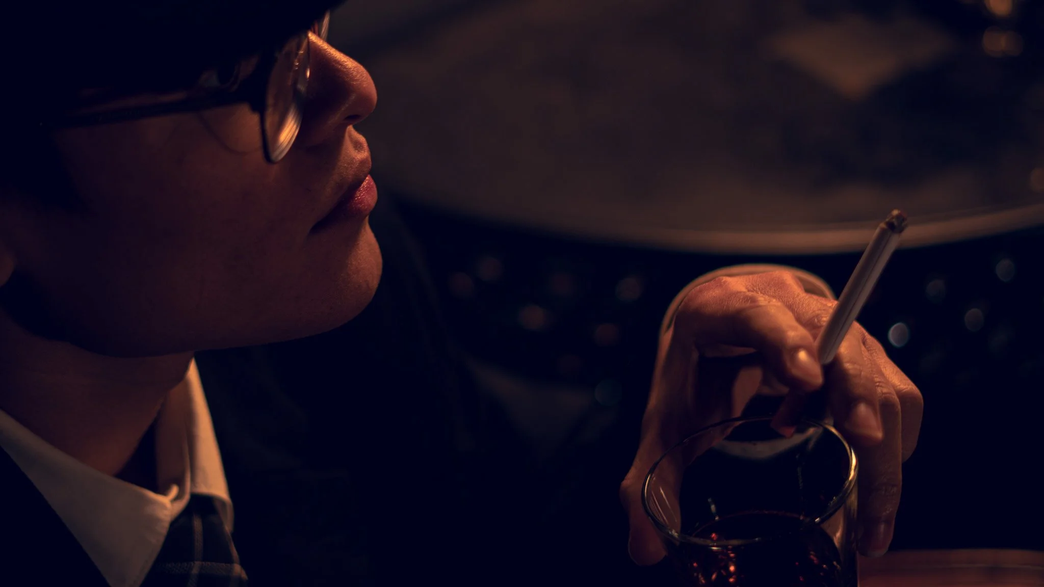 Close-up of a person with glasses holding a drink and smoking a cigarette in a dark setting with warm lighting.