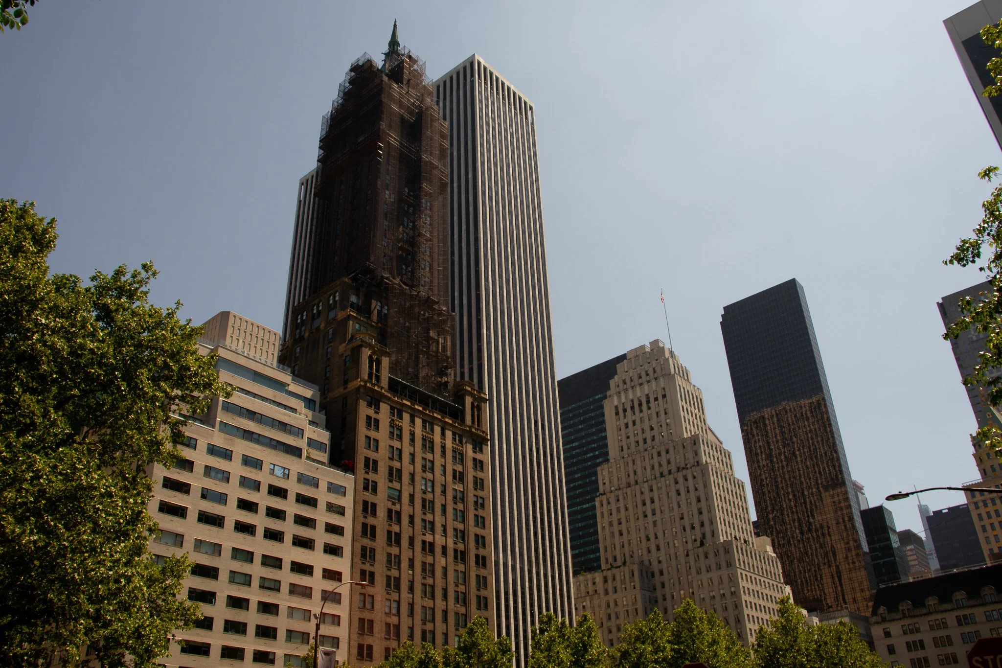 A cityscape with tall skyscrapers, some under construction, set against a clear sky with a few trees in the foreground.