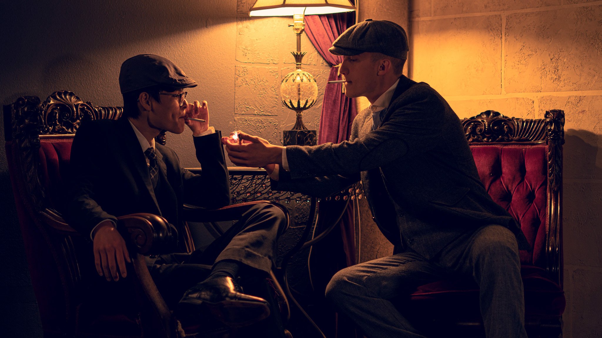 Two men in suits and hats, sitting in a dimly lit room, one man offers a cigarette and lighter to the other.