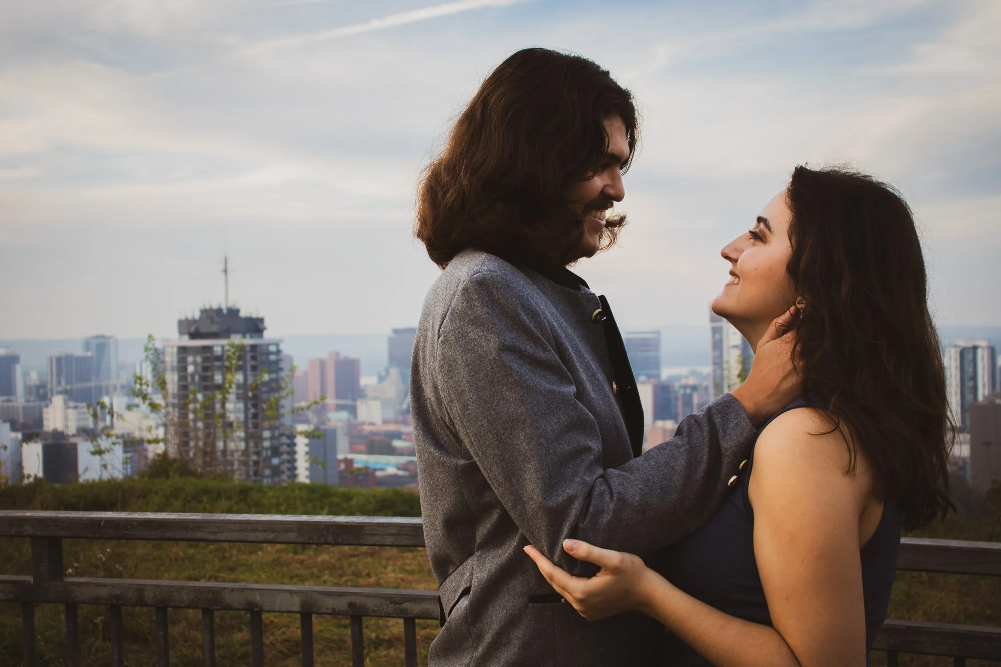A couple is smiling and looking at each other, standing outdoors on a rooftop with a city skyline, including tall buildings and an oil platform, in the background.