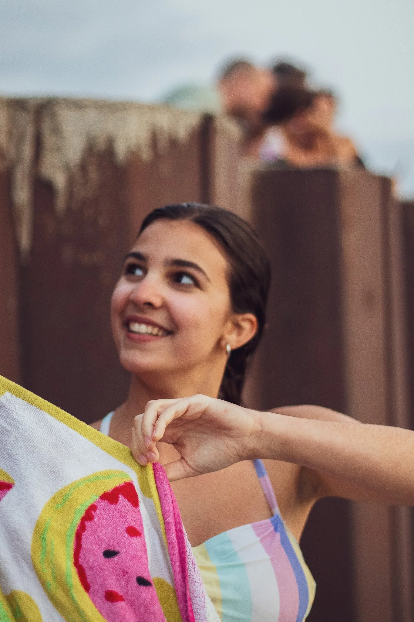 A young woman smiling and looking up, holding a colorful towel with a watermelon pattern, with two people blurred in the background.