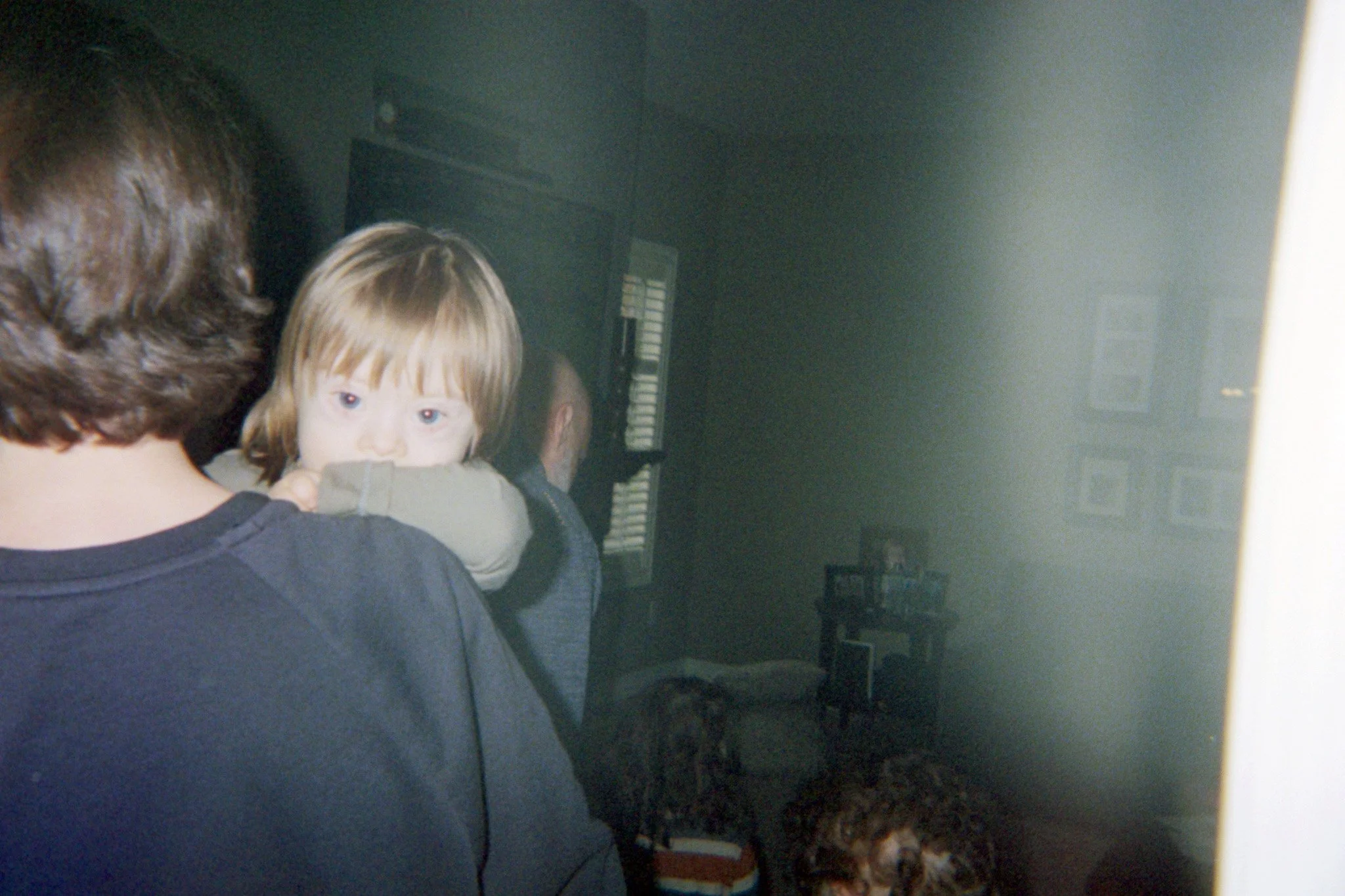 A man with brown hair seen from behind, holding a young boy with reddish-brown hair, who is looking directly at the camera with wide eyes, in a dimly lit room.