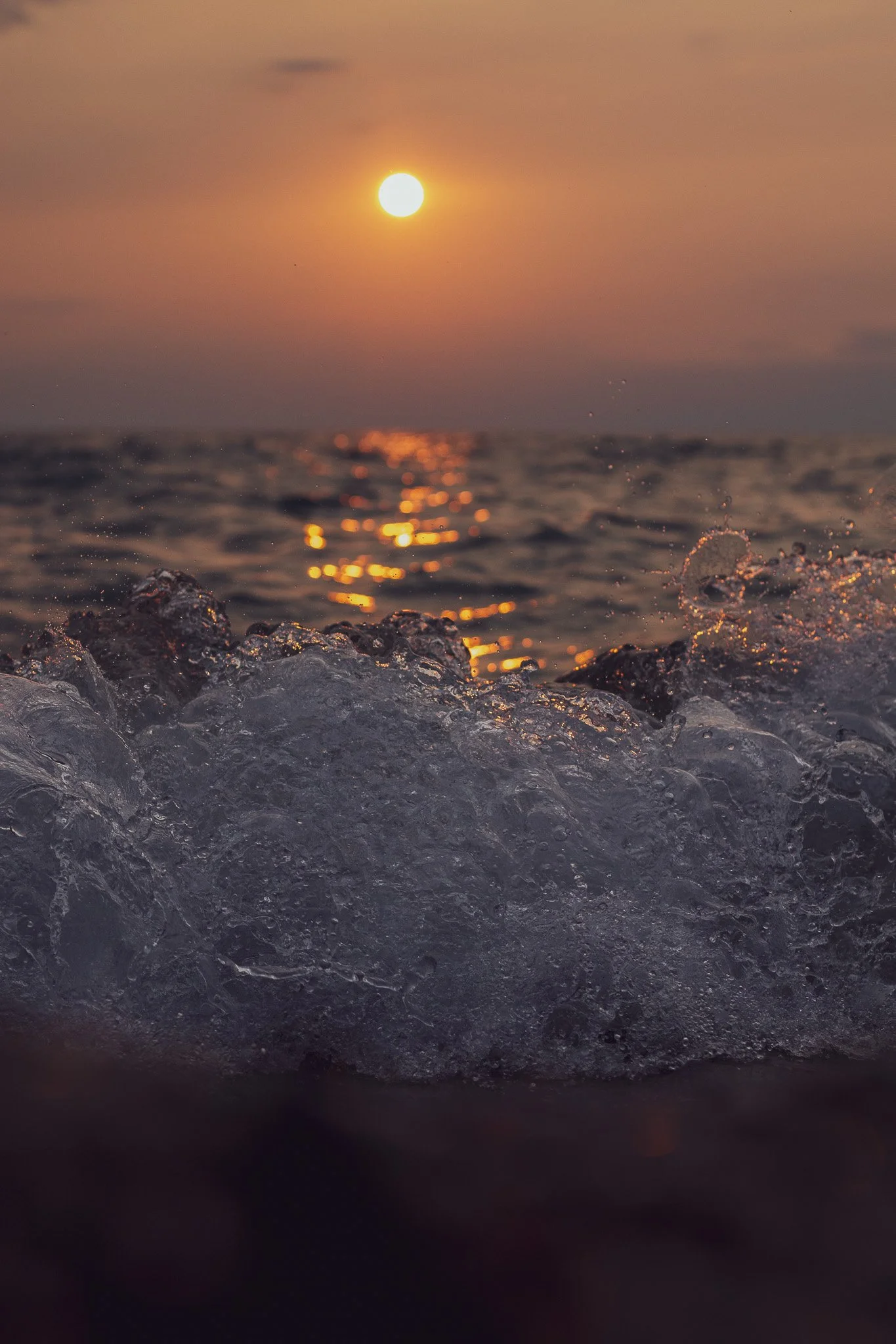 A photo of the ocean at sunset with the sun visible in the sky, reflecting on the water, and waves crashing at the shoreline.