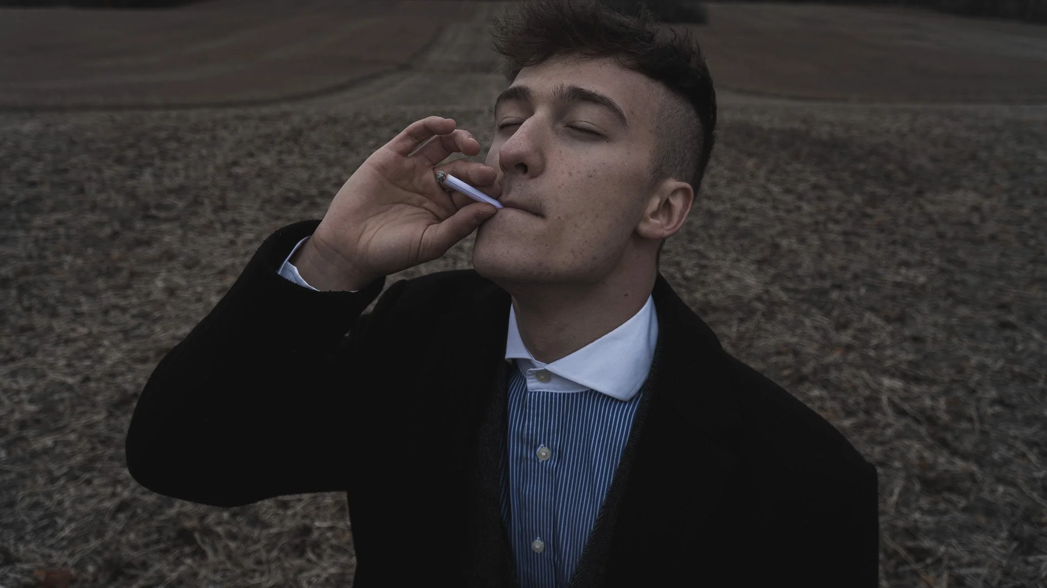 A young man with short dark hair, dressed in a black jacket and a blue striped shirt, is smoking a cigarette outdoors on a cloudy day, with a blurred background of grass or bare ground.