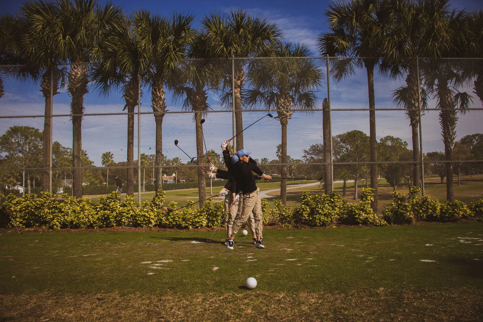 People practicing golf on a driving range with tall palm trees and a chain-link fence in the background under a partly cloudy sky.