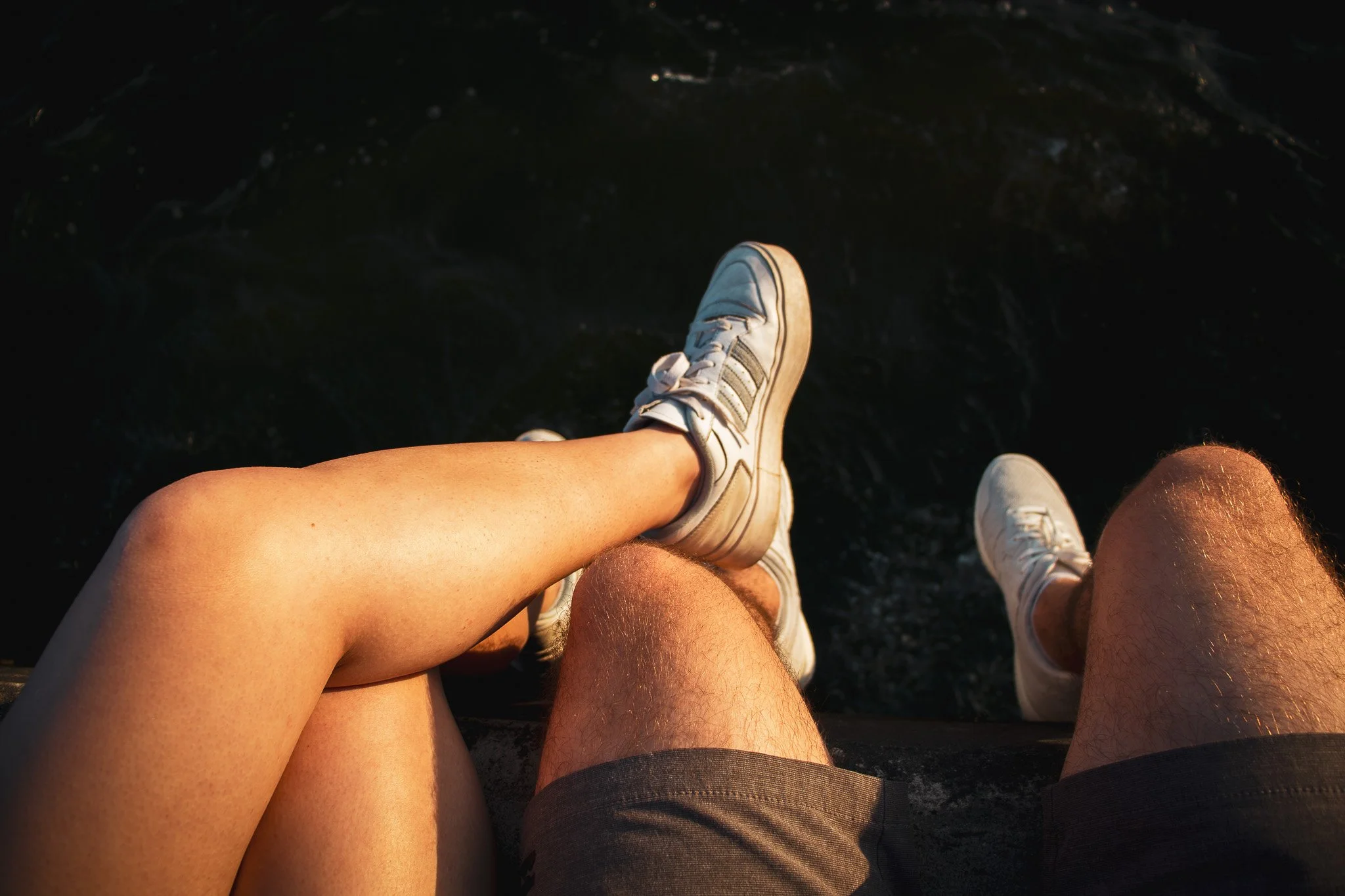 Two pairs of legs, one with blonde hair, wearing white sneakers and shorts, sitting with their legs extended downward over what appears to be a dark water body.
