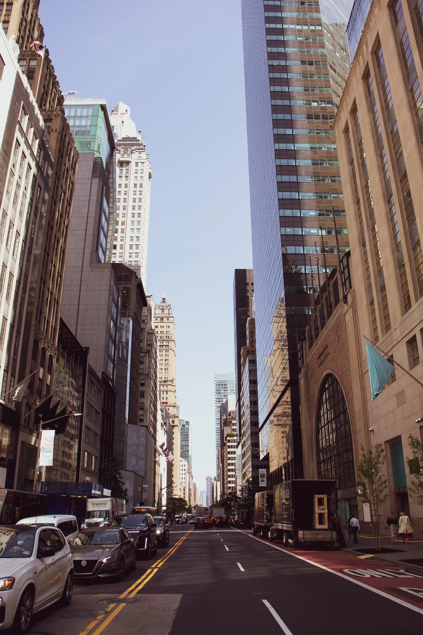 City street with tall skyscrapers on both sides, cars parked and driving, and pedestrians walking along the sidewalk.
