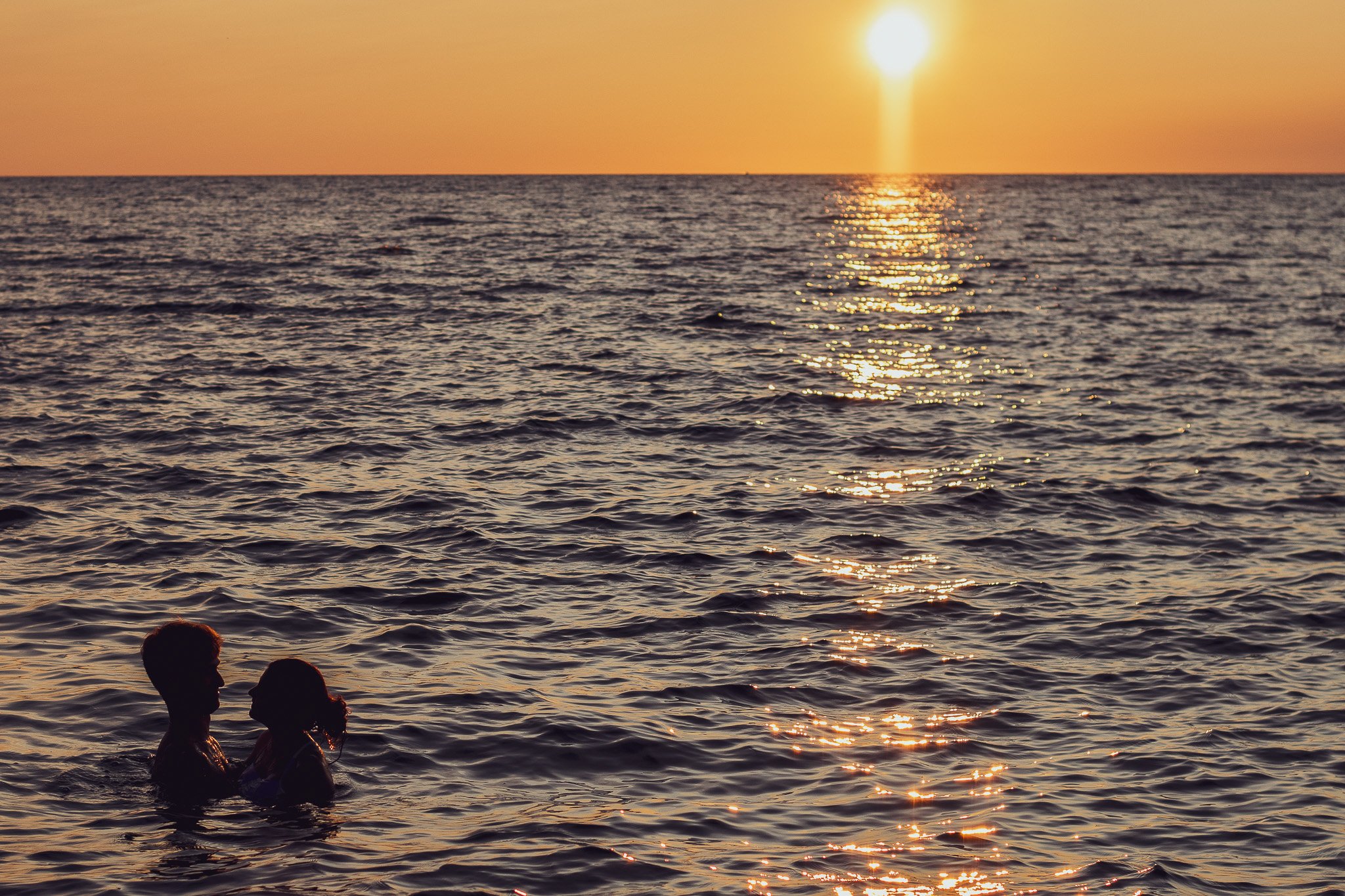 A sunset over the ocean with reflections on the water. Silhouettes of two people standing in the water are visible in the foreground.