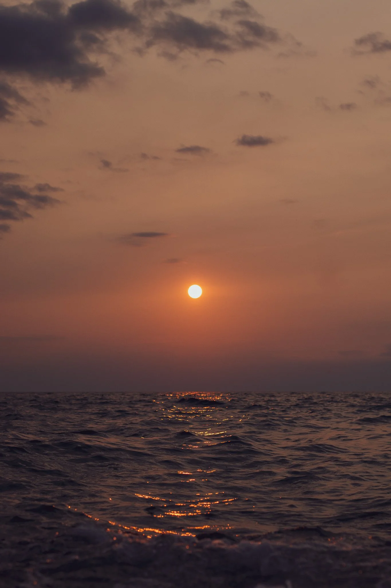 A sunset over the ocean with the sun near the horizon, casting an orange glow and reflecting on the water, with clouds in the sky.