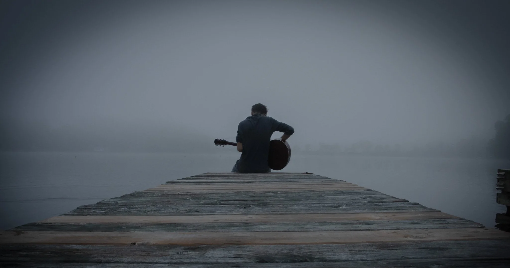 A person sitting on a wooden dock by a foggy lake, holding a guitar and facing away from the camera.