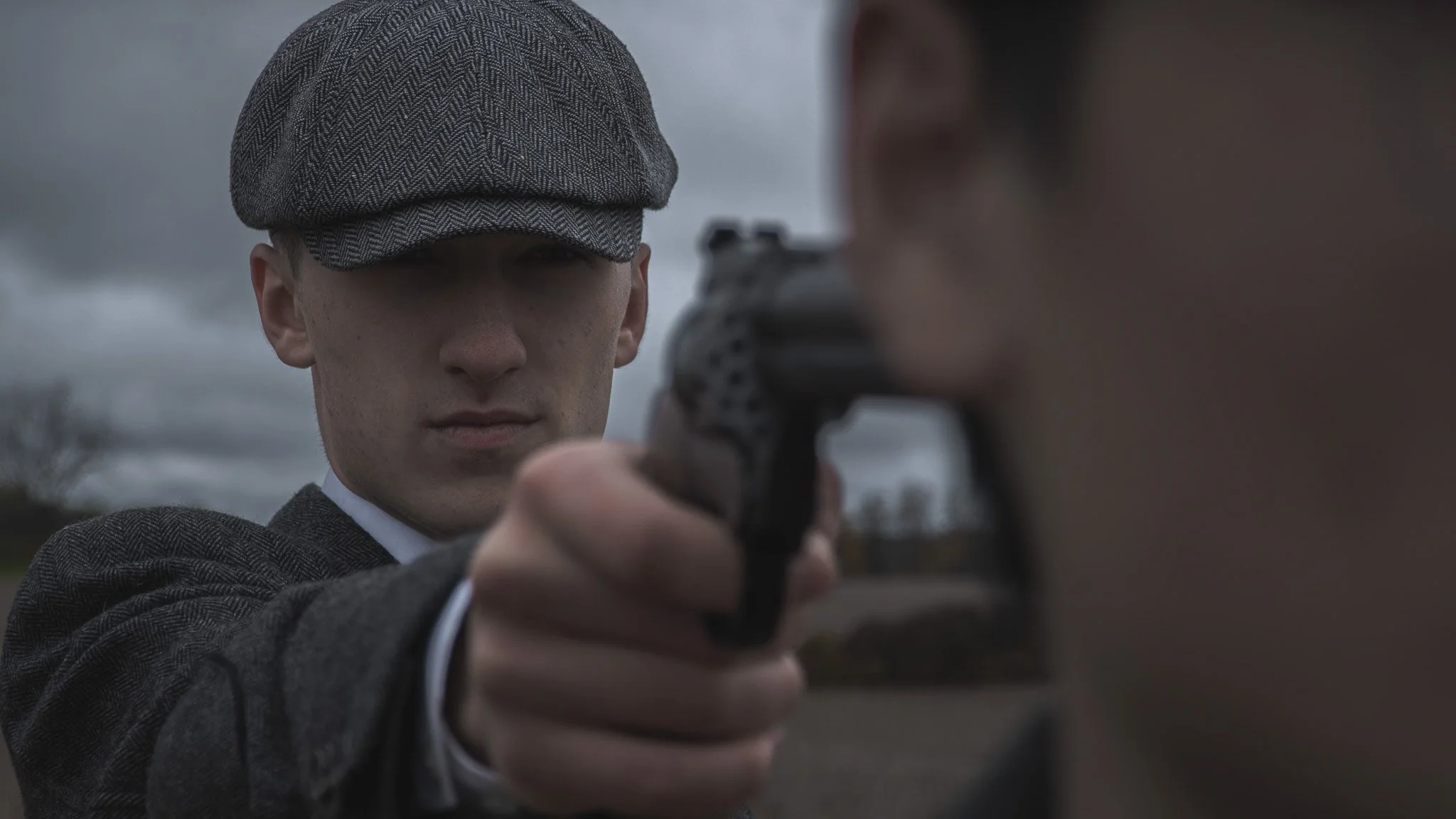 A man wearing a gray herringbone flat cap and a matching gray tweed coat is pointing a gun directly at the camera. The background is cloudy and overcast.