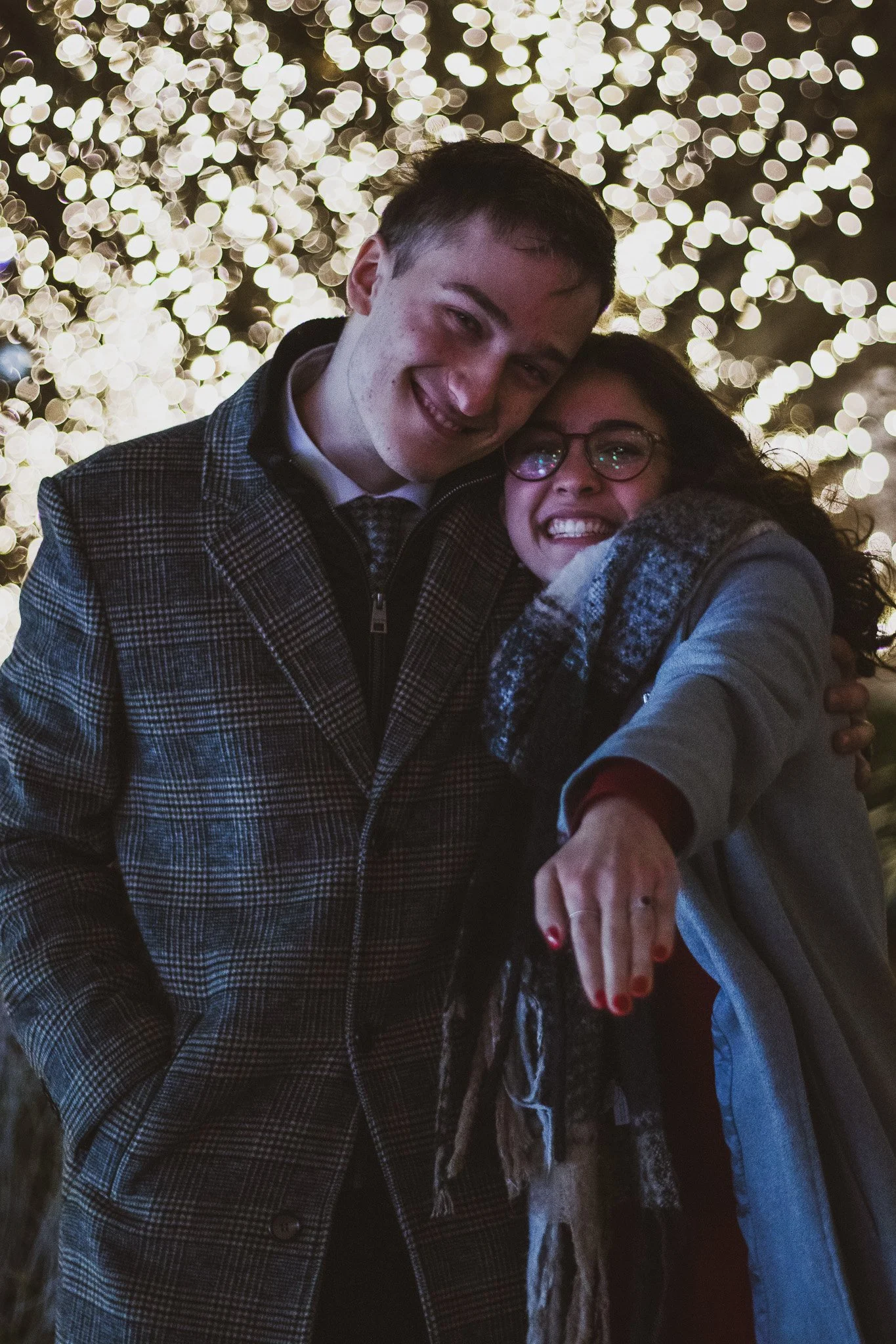 Two people smiling and taking a selfie in front of a background of blurry, glowing Christmas lights.
