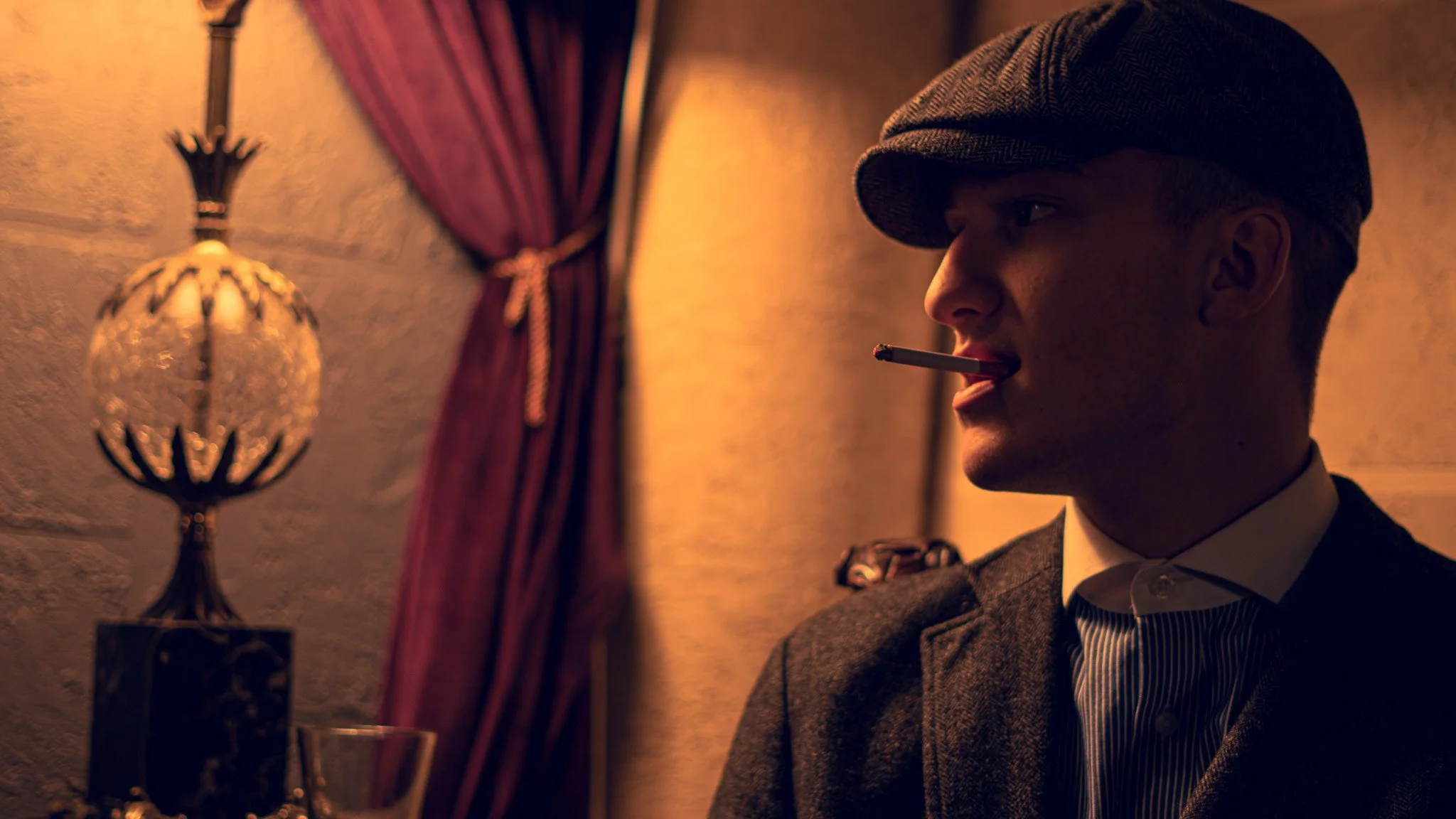 A man in vintage style clothing with a flat cap chewing on a cigarette, standing in a dimly lit room with a decorative lamp and red curtain.