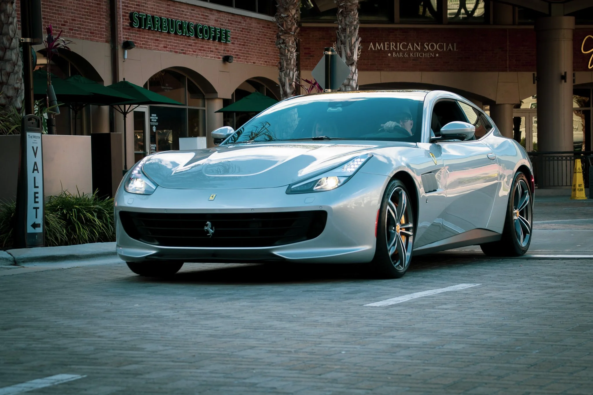 A white Ferrari parked on the street outside a restaurant called American Social Bar & Kitchen with a Starbucks Coffee shop in the background.