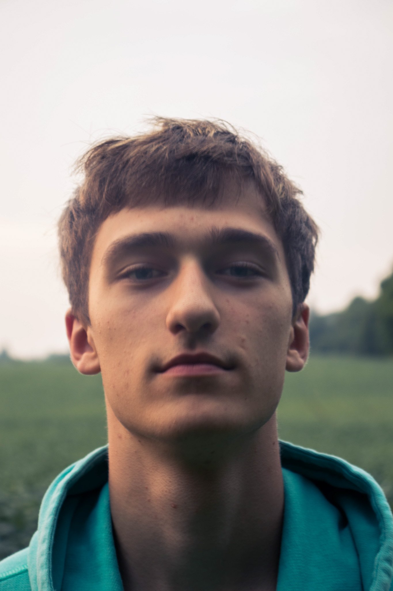 Close-up of a young man's face with short brown hair, wearing a teal hoodie, outdoors with a blurred natural background.