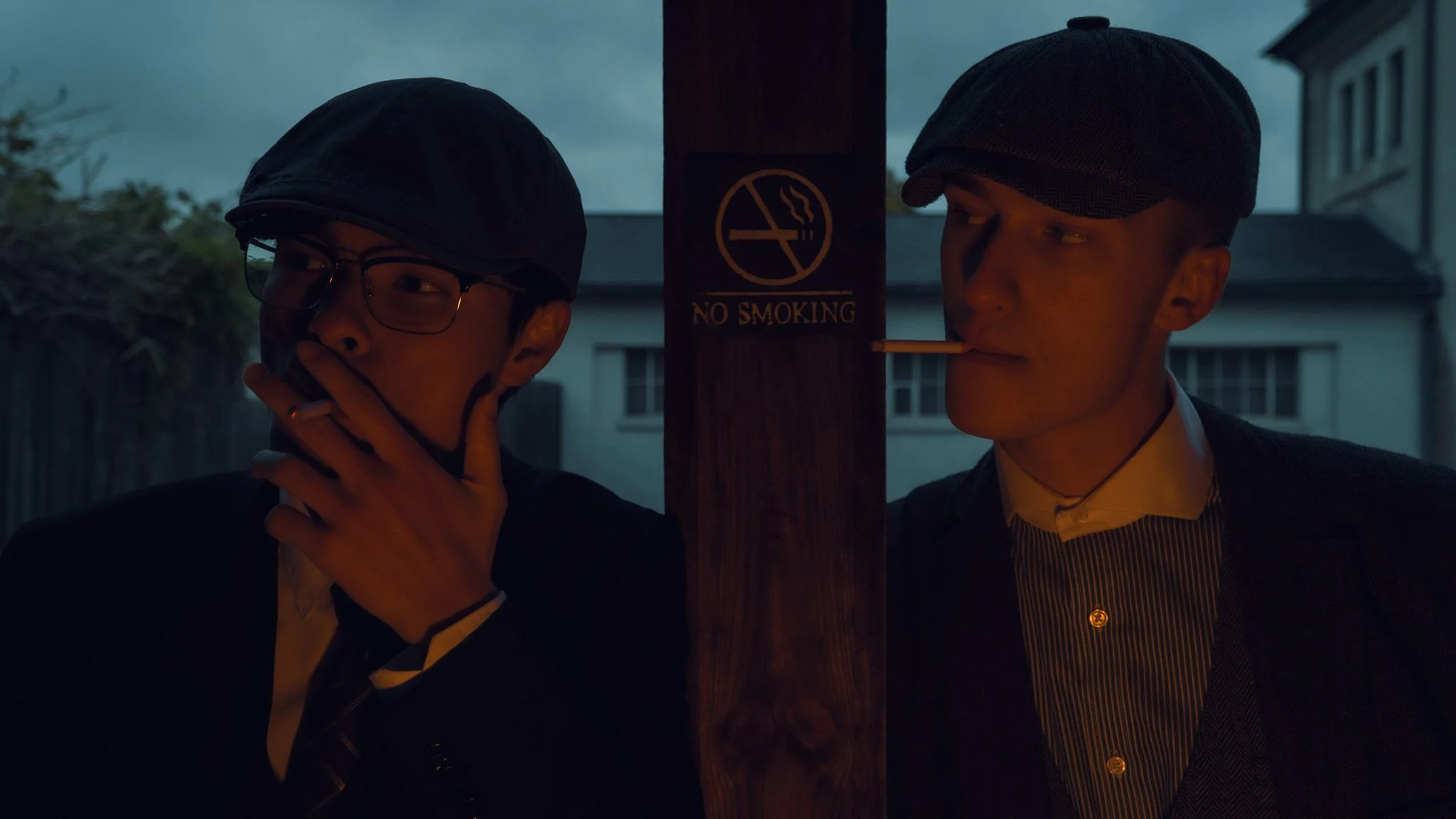 Two young men with hats and glasses smoking cigarettes while standing near a 'No Smoking' sign on a wooden post outdoors at dusk.