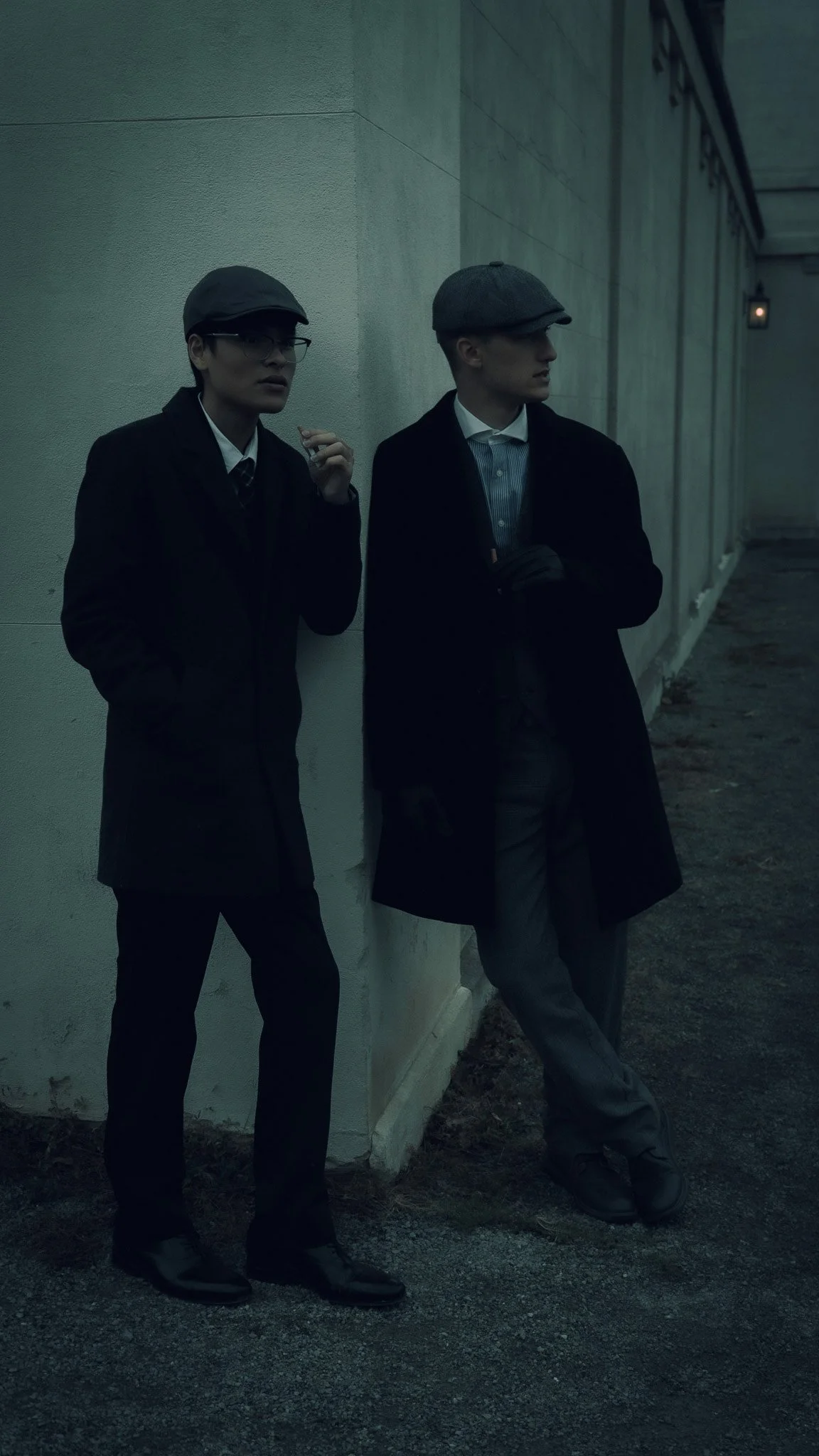 Two individuals dressed in vintage-inspired clothing, standing outside against a concrete wall. They are wearing dark coats, hats, and glasses, with a moody, subdued atmosphere.