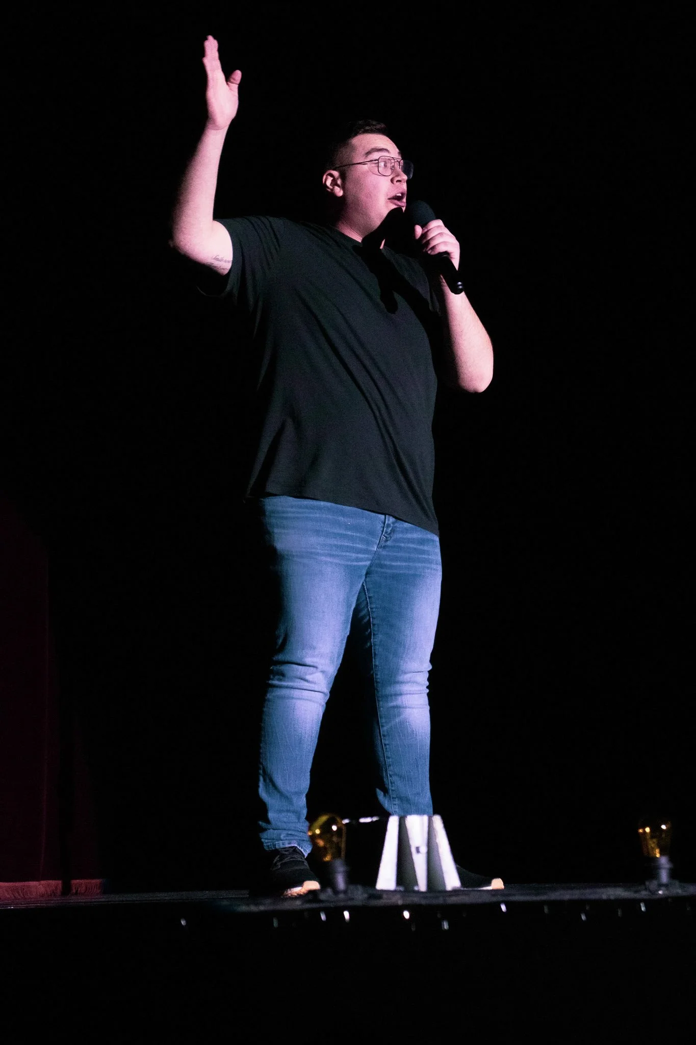 A person standing on a stage, holding a microphone in one hand and raising the other hand, against a dark background.