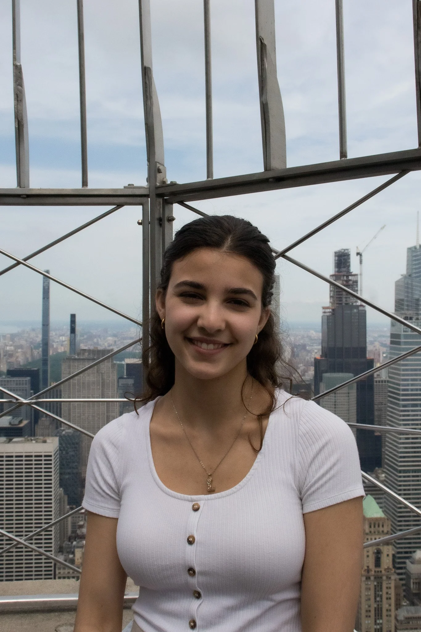 A smiling woman with dark hair tied back, wearing earrings, a white button-up shirt with three visible buttons, and a necklace, standing on a rooftop with a city skyline in the background.