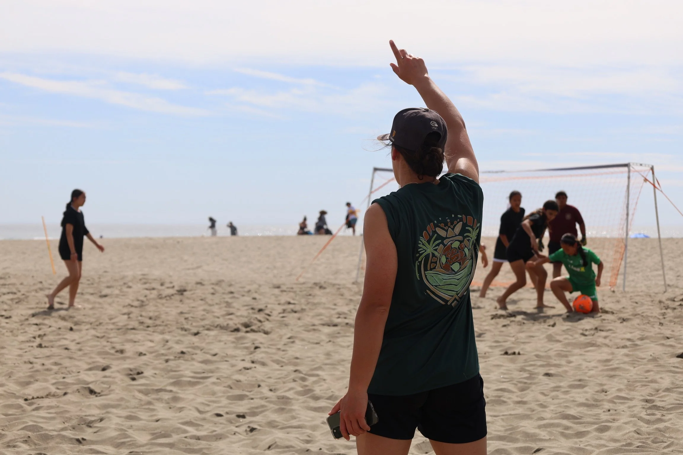 A woman in a black cap and black shirt with a colorful design on the back is standing on a sandy beach, pointing upward with her right arm. In the background, there are people playing volleyball, some sitting and others standing near the net, under a sky with some clouds.