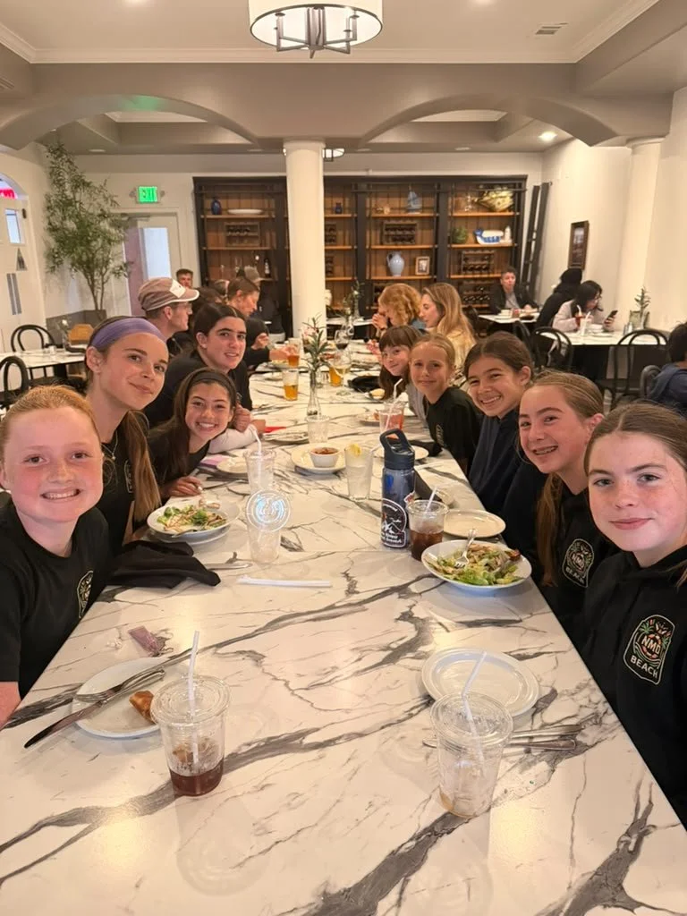 Group of children sitting at a long marble table in a restaurant or dining room, smiling, with plates of food, drinks, and napkins, with a background of wooden shelves and white columns.