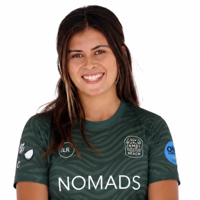 A woman with long dark hair, smiling, wearing a green Nomads sports jersey with various patches and logos, against a white background.