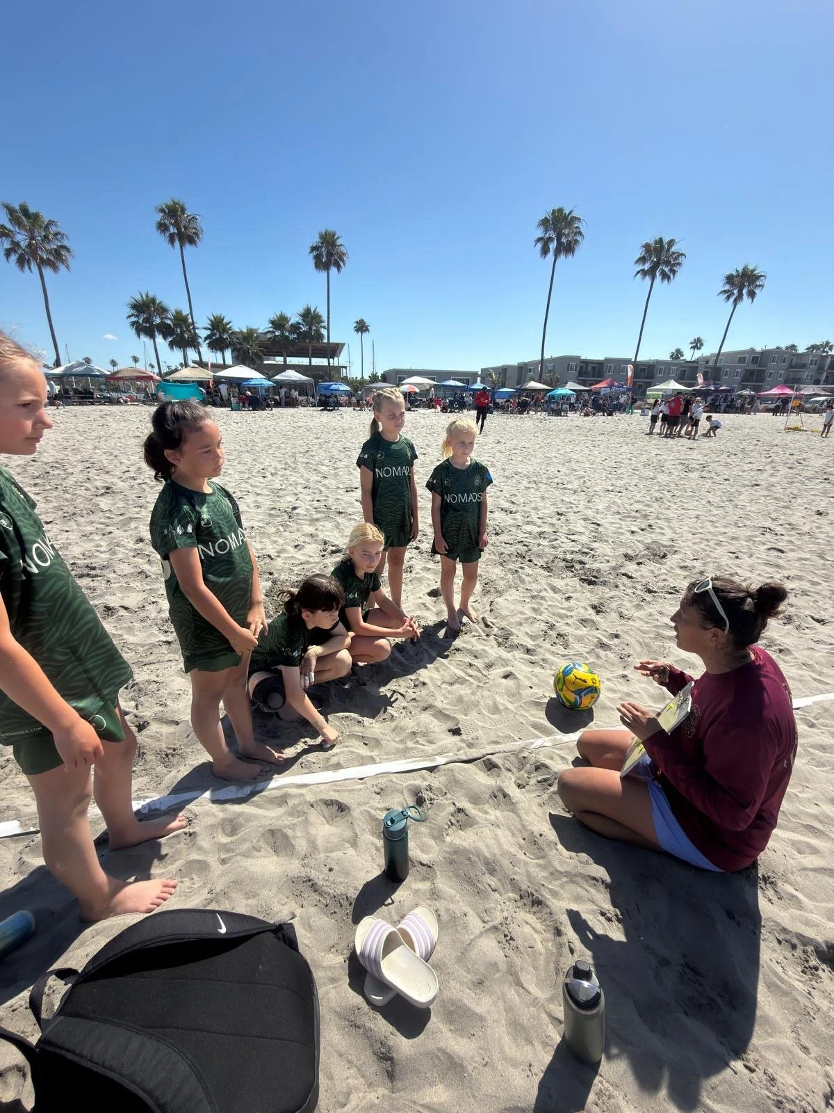 A group of children in green sports uniforms gathered on a sandy beach, listening to a woman sitting on the sand. The woman appears to be giving instructions or coaching, with a soccer ball nearby. In the background, there are palm trees, umbrellas, and people in front of beachside buildings, under a clear blue sky.