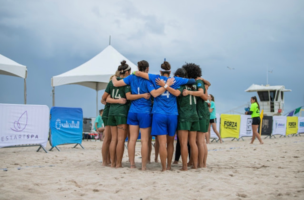 A group of women on a beach, wearing sports uniforms, found hugging in a circle.