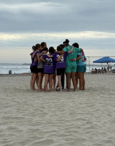 Group of women and girls in a huddle on a beach, some wearing sports jerseys, with ocean and beach tents in the background.
