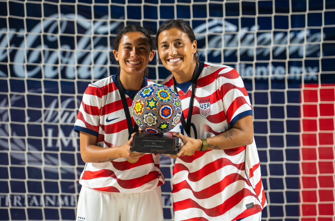 Two female soccer players in striped USA jerseys holding a colorful soccer ball and a trophy, standing in front of a goal net.