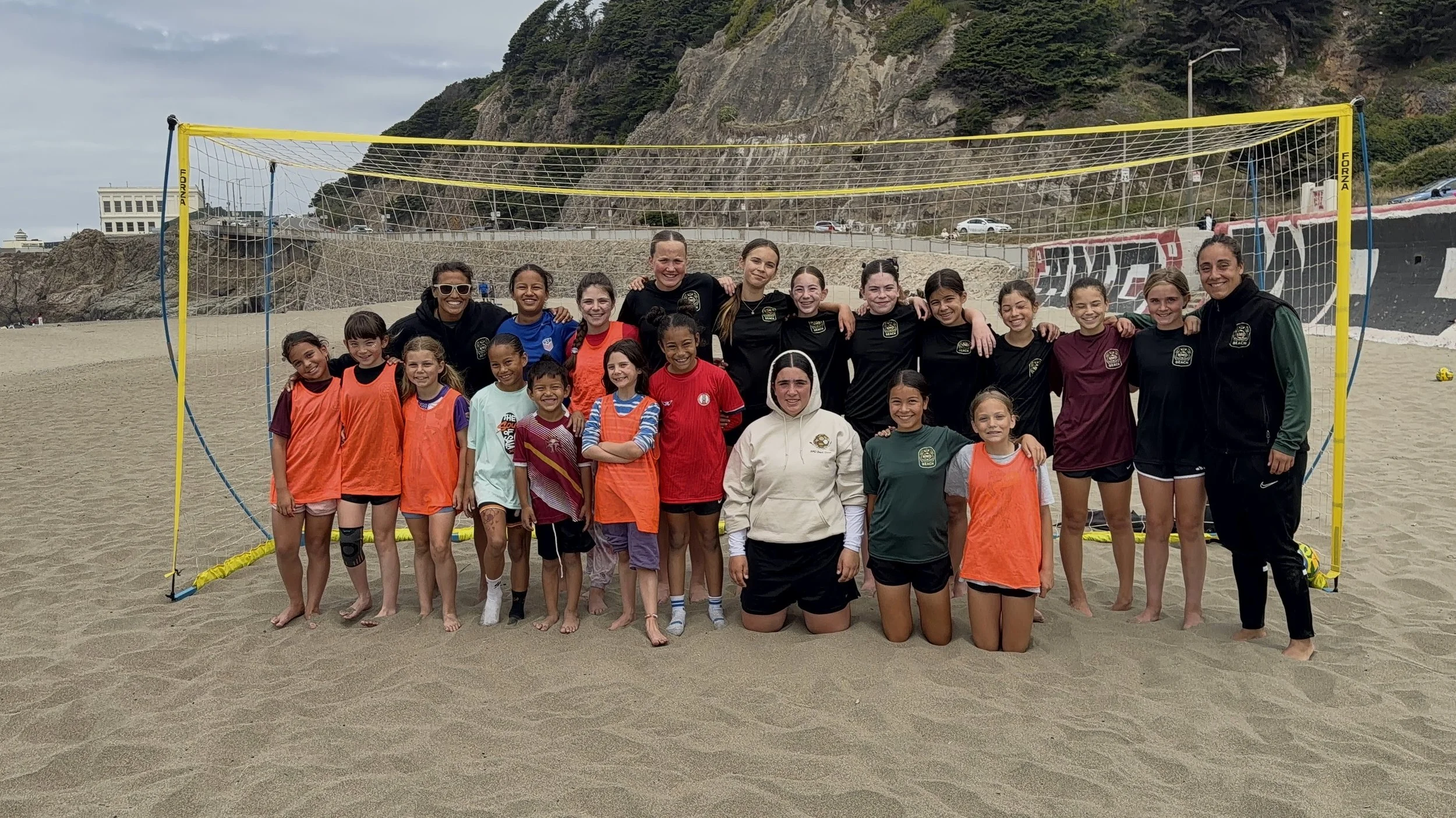 A group of young girls and women, some wearing sports gear, standing together on a sandy beach in front of a yellow soccer goal. The group appears to be part of a soccer team or camp, with a scenic backdrop of cliffs, a street with cars, and cloudy sky.