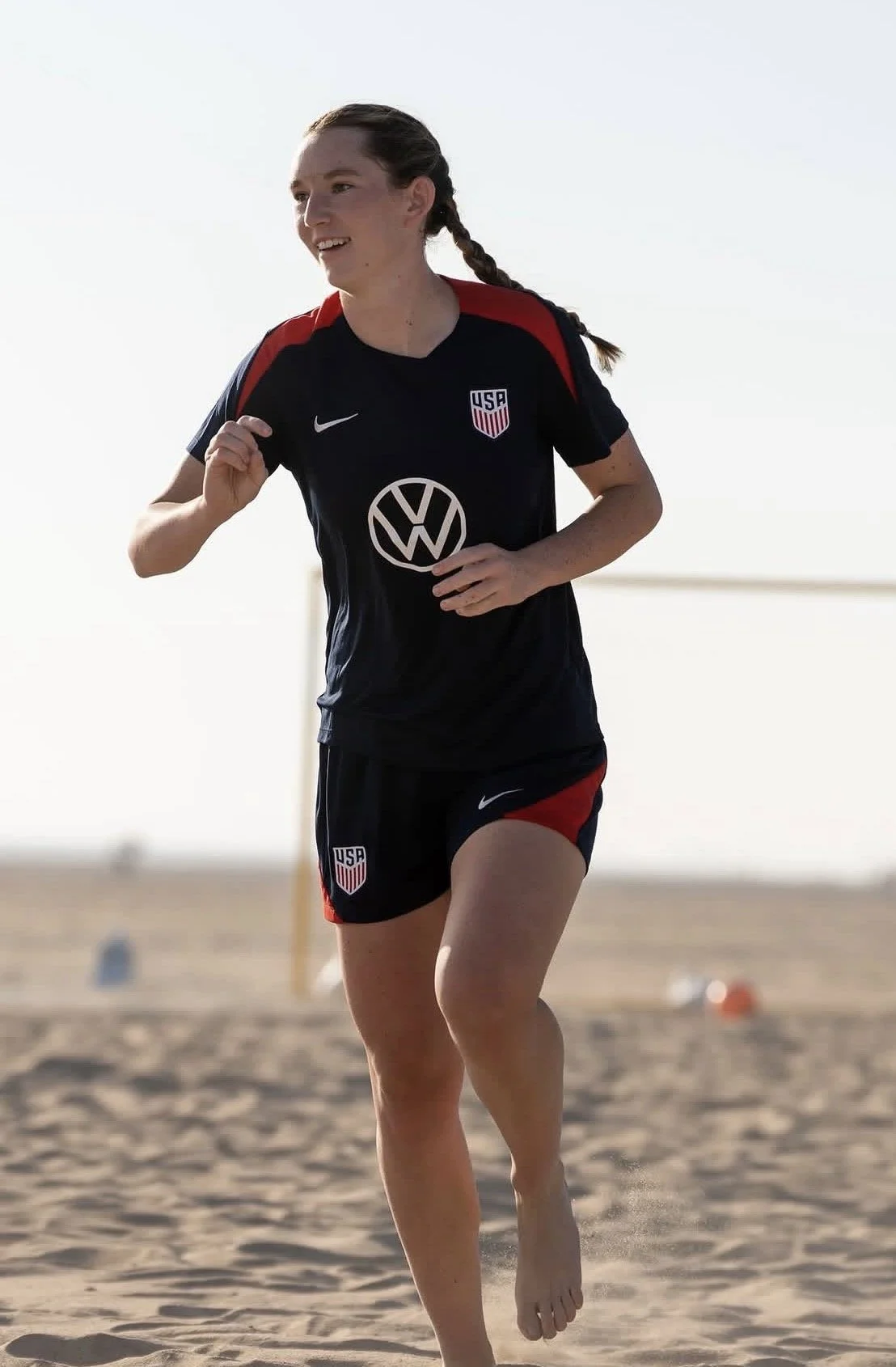 A woman running on the beach, wearing a USA soccer uniform with a black and red jersey and matching shorts.