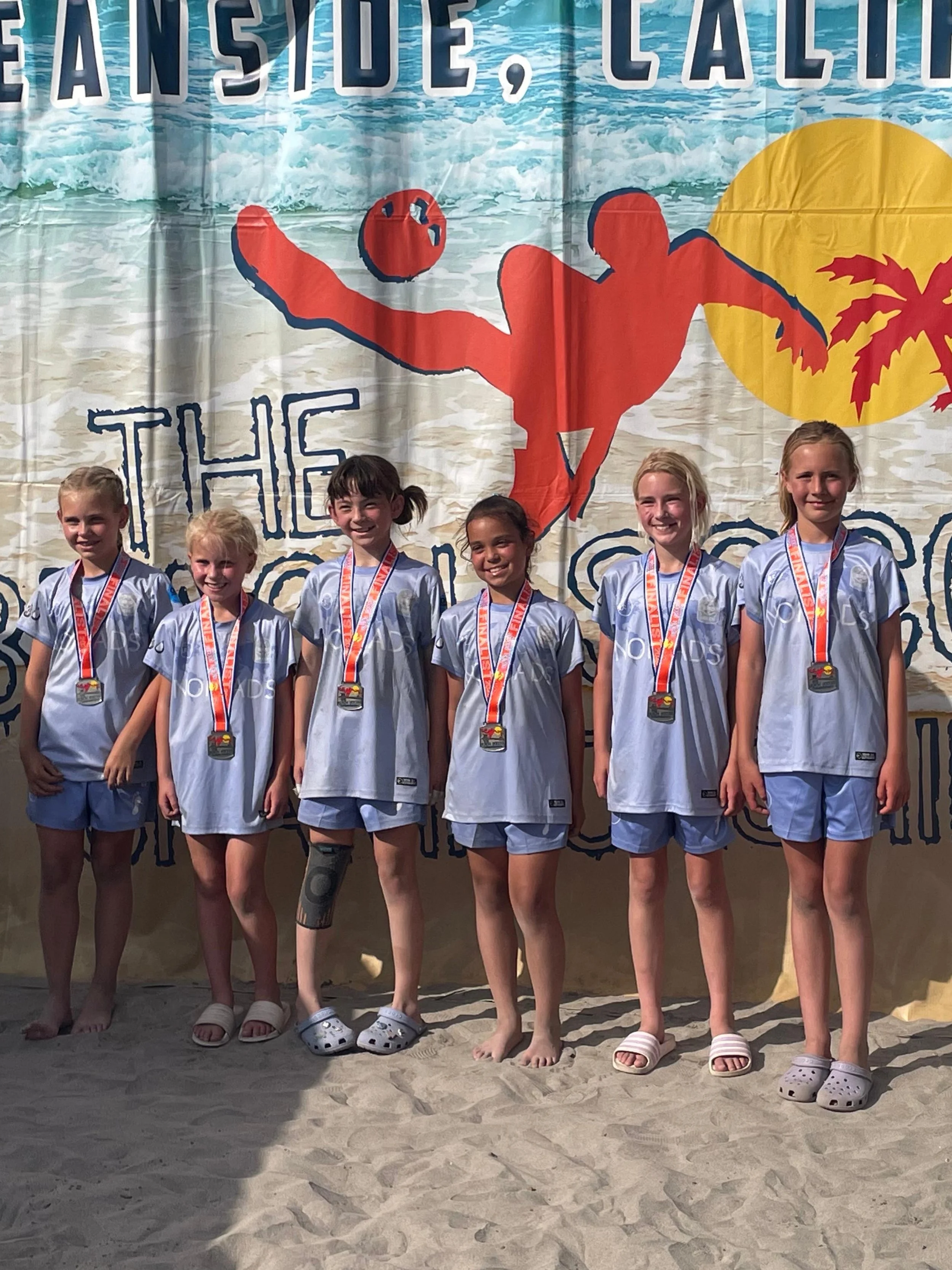 Six young girls standing on a sandy beach in front of a large ocean-themed banner. They are wearing matching gray sports jerseys and medals around their necks. Some are wearing different types of footwear, and one girl has a medical knee brace.