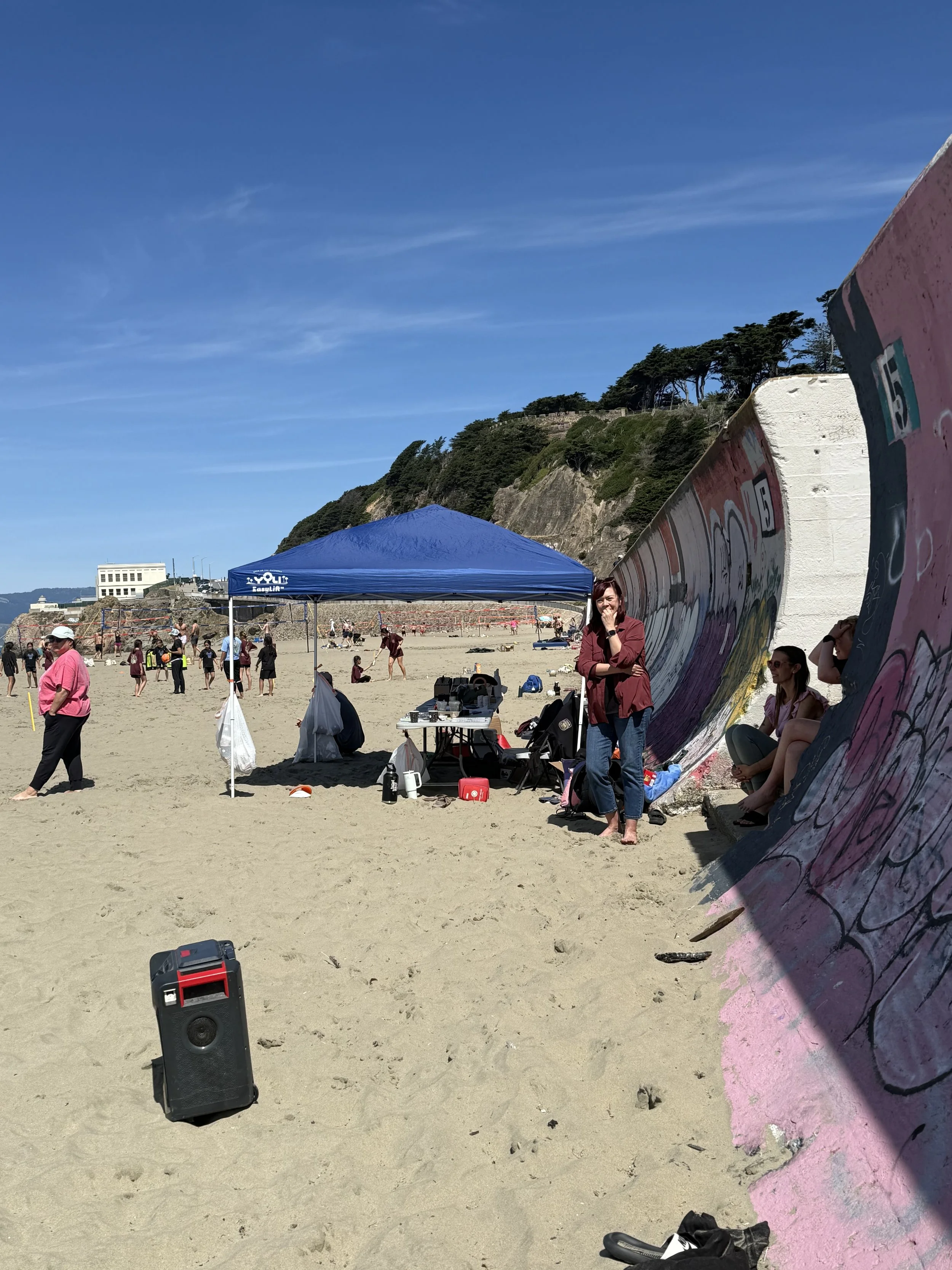 Beach scene with people, a blue canopy, and a large painted concrete structure with graffiti. A woman stands near the structure, and others are sitting or walking on the sand. There is a speaker in the foreground and hills with trees in the background.