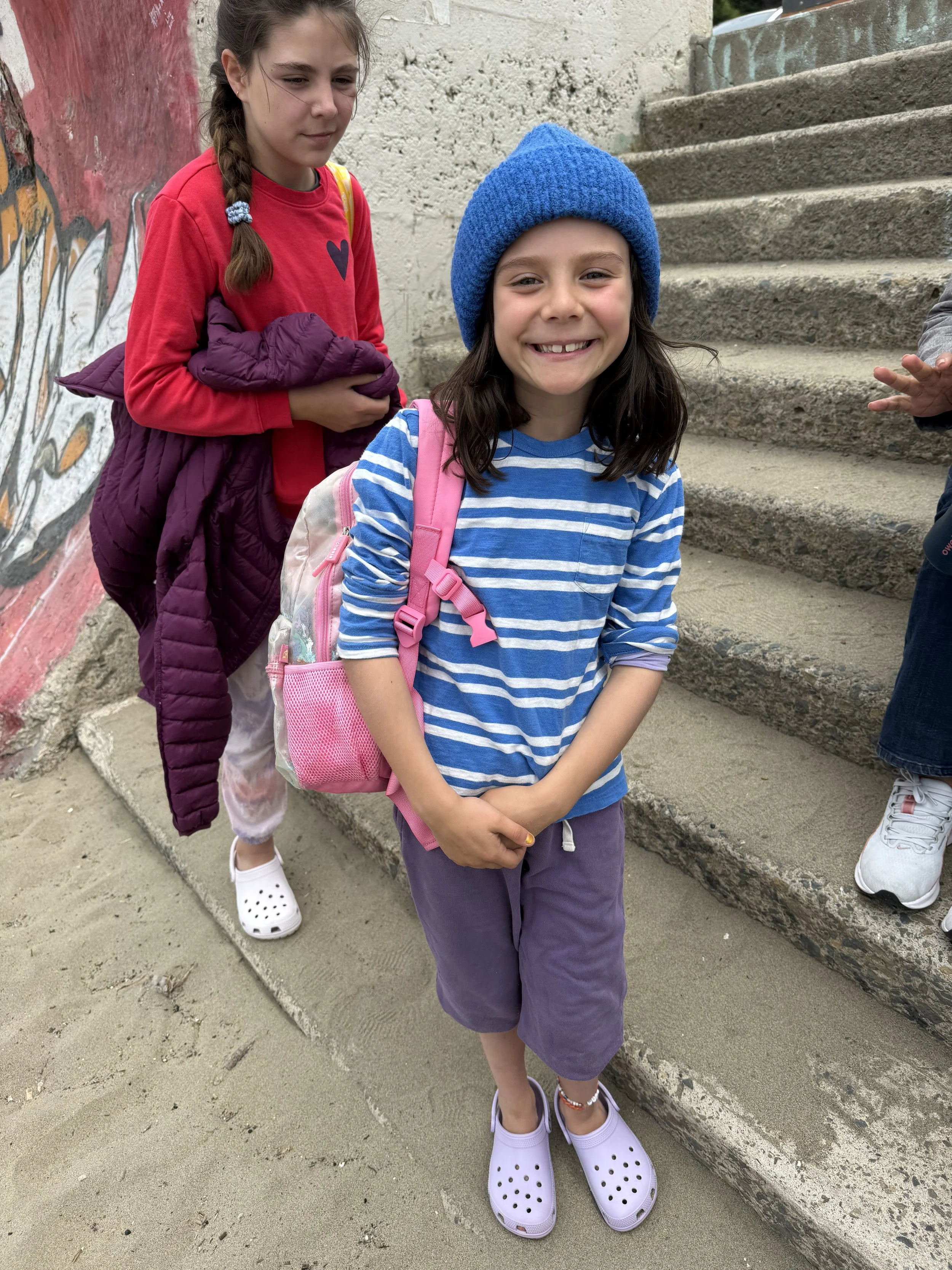 Two young girls standing on concrete steps outside. One girl in the foreground is smiling, wearing a blue beanie, striped blue and white shirt, purple pants, and white Crocs. The girl behind her is looking down, dressed in a red shirt with a heart on it, white pants, and holding a purple jacket. They are next to a wall with graffiti.