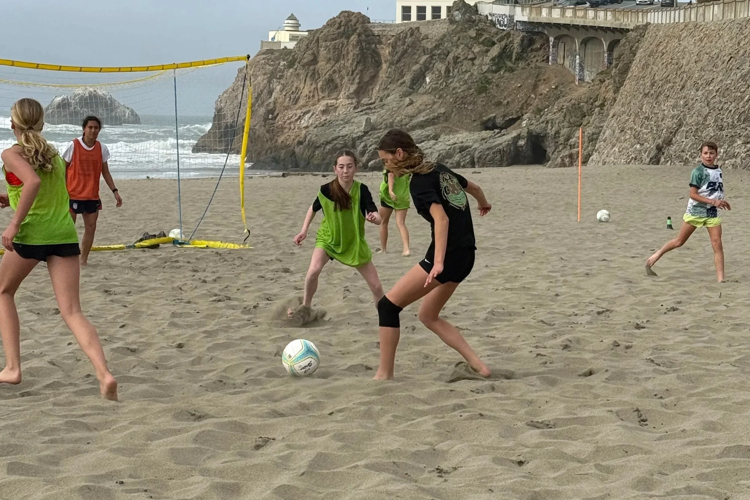 People playing beach volleyball on the sand near the ocean with rocky cliffs and buildings in the background.
