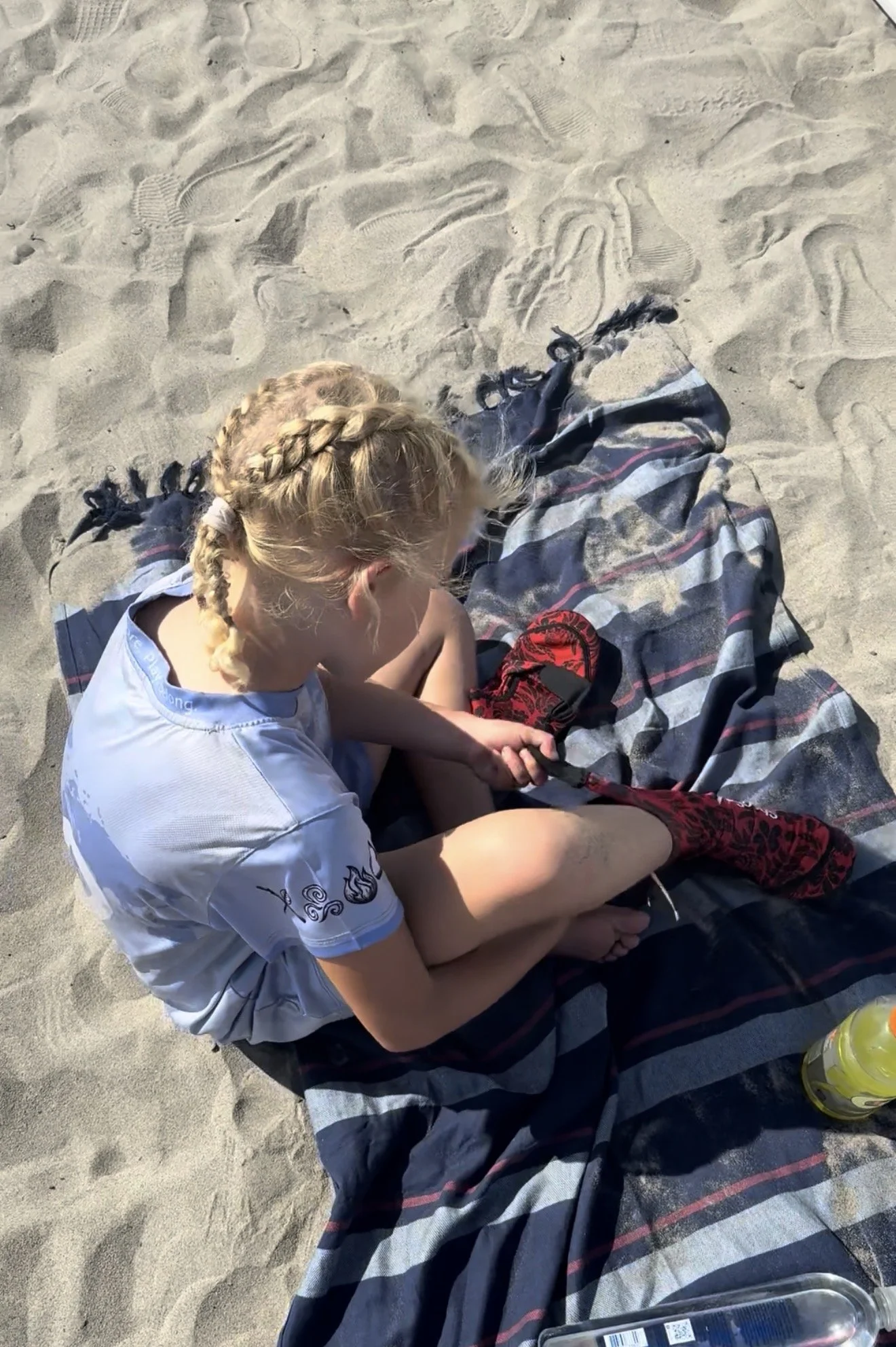 A young girl with blonde hair in a braid is sitting on a striped blanket on the sand at the beach. She is wearing a white t-shirt with black designs on the sleeve and is holding a red and black item. There are footprints and ridges in the sand around her, and a clear plastic water bottle is partially visible in the lower right corner.