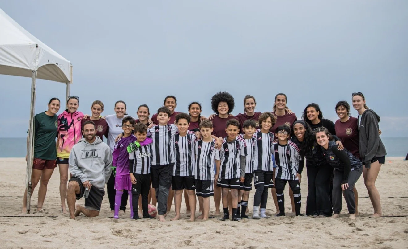 Group of children and adults on the beach, posing for a photo, with some wearing sports jerseys and others in casual clothes, under a cloudy sky.