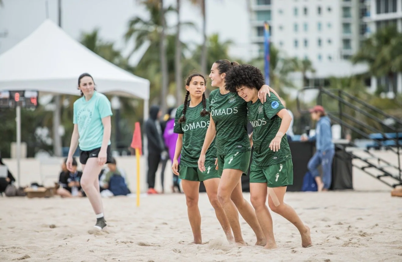 Three women in green sports uniforms walking on sandy beach, with one supporting another by the shoulders, and a fourth woman in a light blue shirt and black shorts in the background.